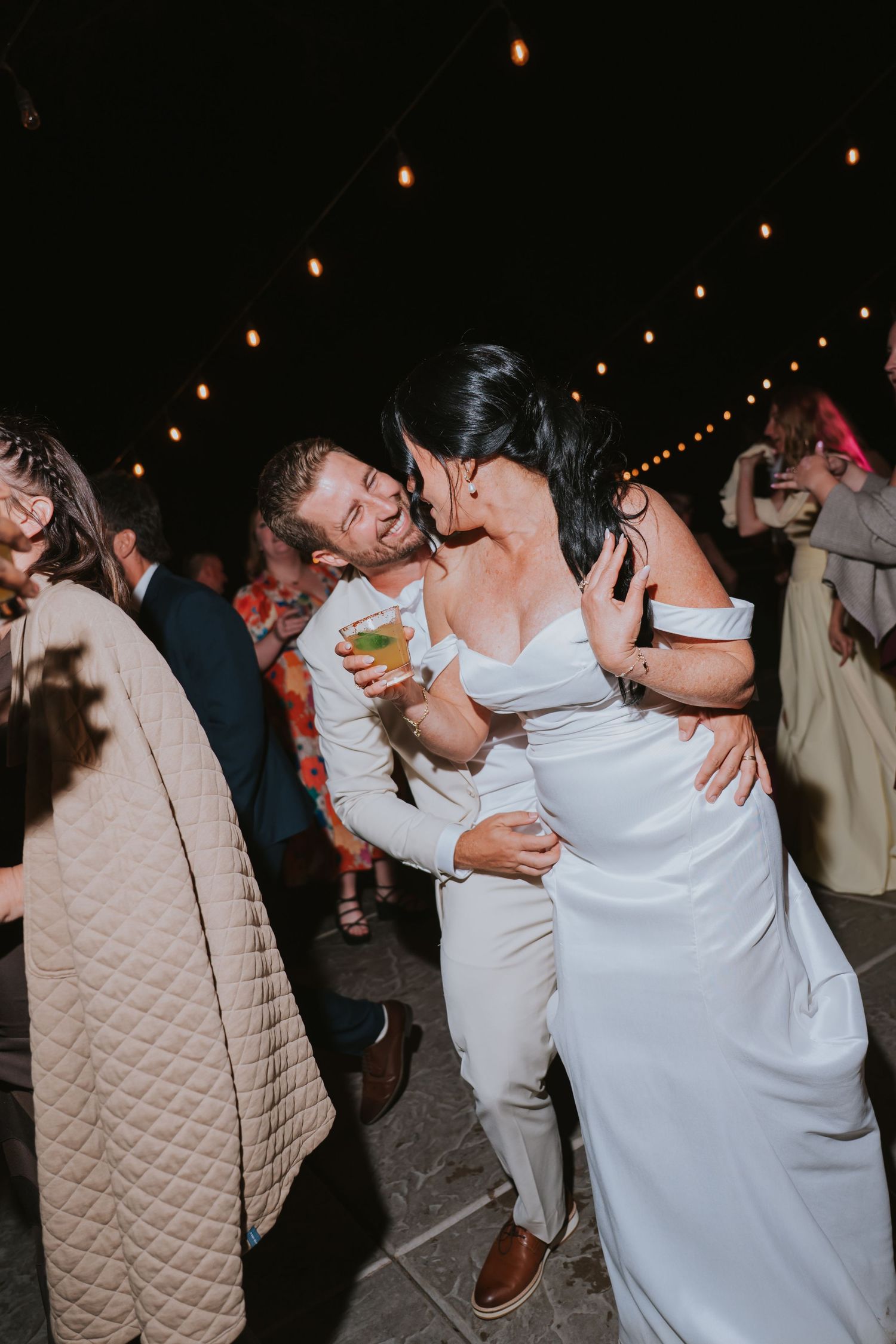 Bride and groom dancing joyfully at outdoor wedding reception under string lights at night.