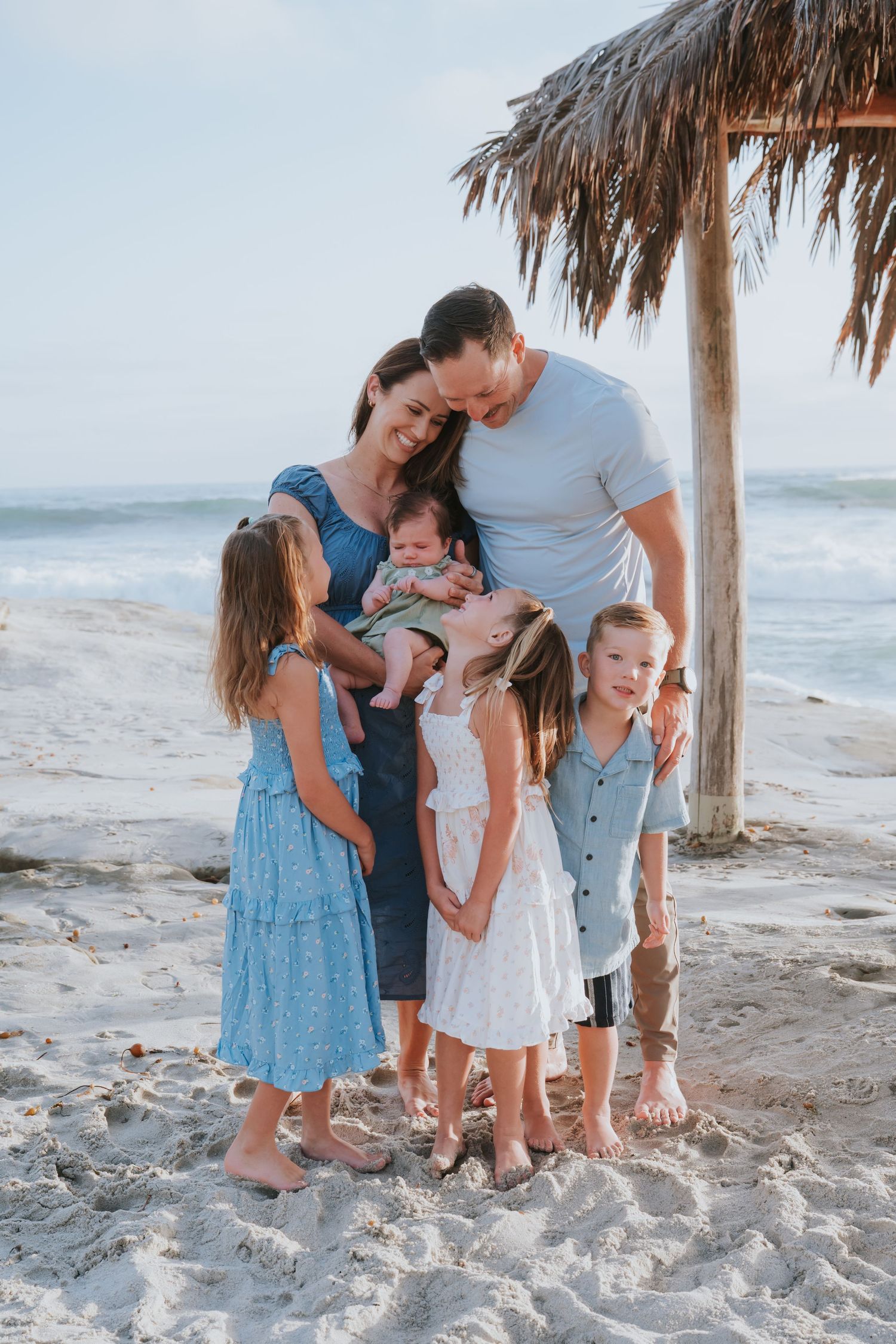 A happy family of six poses together under a thatched palm umbrella on a sandy beach with ocean waves.