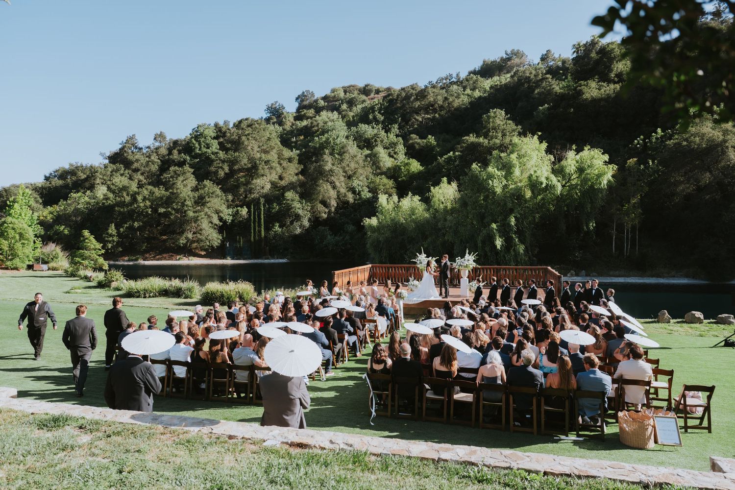 Outdoor wedding ceremony on green lawn near a lake, surrounded by lush trees, with guests seated in wooden chairs.