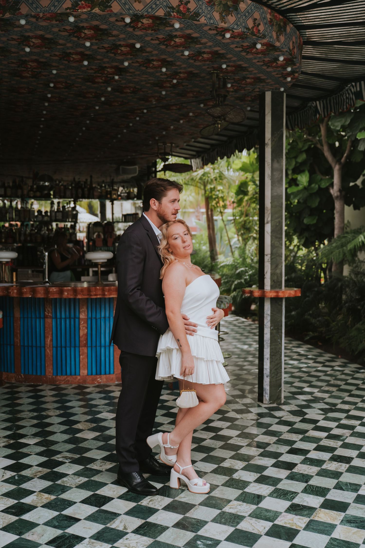 Groom embraces bride from behind on a green and white checkered floor beneath a carousel bar.