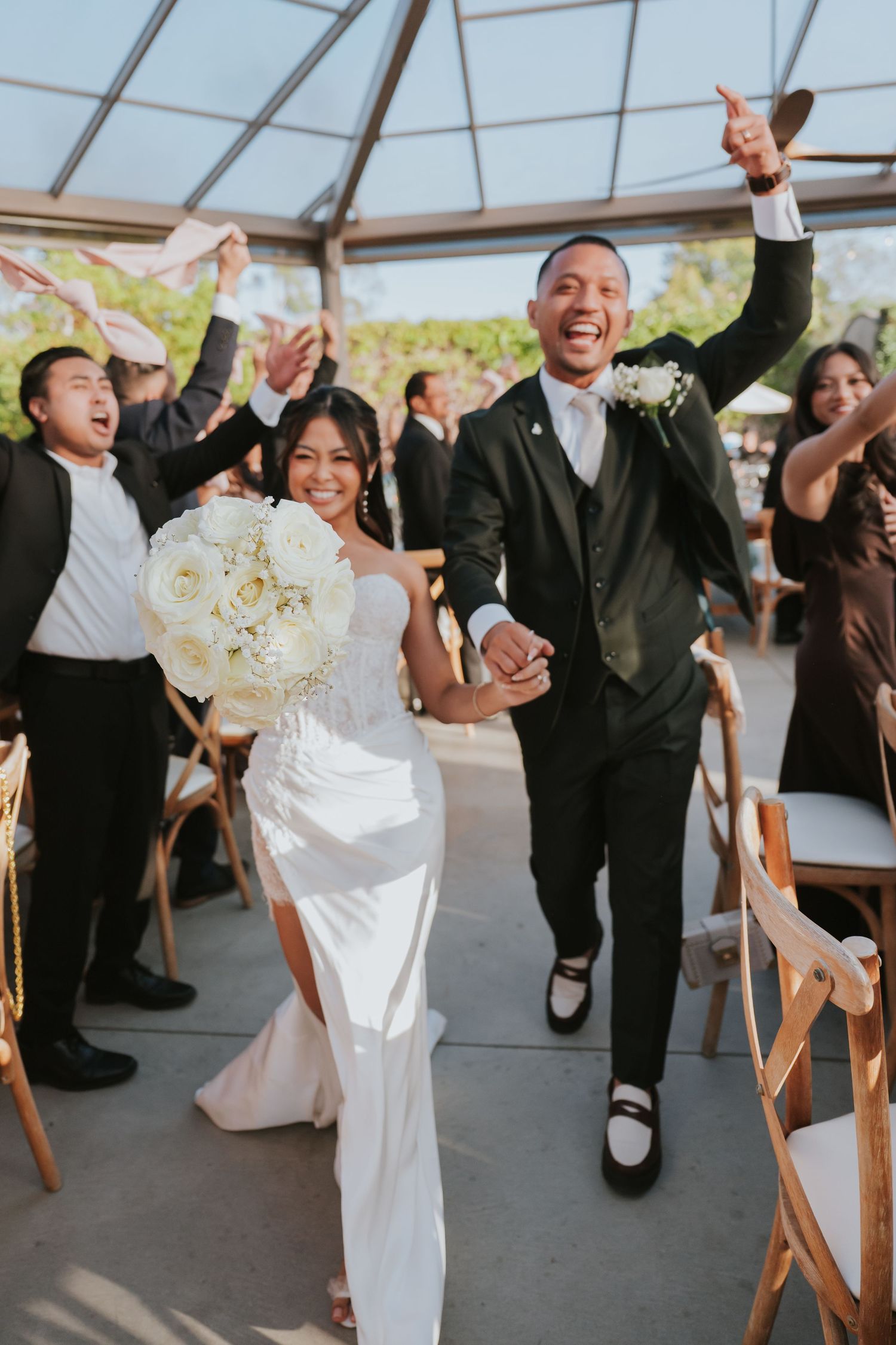 Newlyweds joyfully exit their wedding ceremony under a glass ceiling while guests celebrate with raised hands.