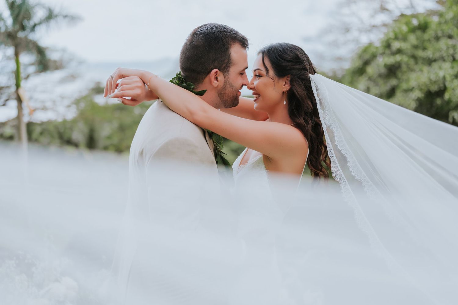 Romantic wedding couple shares an intimate moment with flowing veil against cloudy sky.