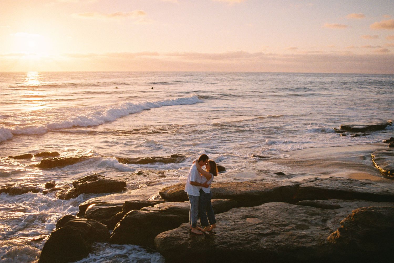 Couple embracing on rocky shore at golden sunset with ocean waves, romantic beach engagement photo.