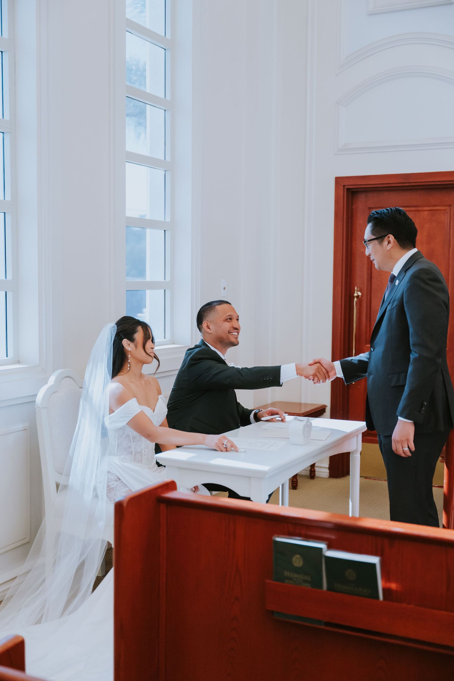 A couple in wedding attire sits at a table while shaking hands with someone in a suit during a formal ceremony.