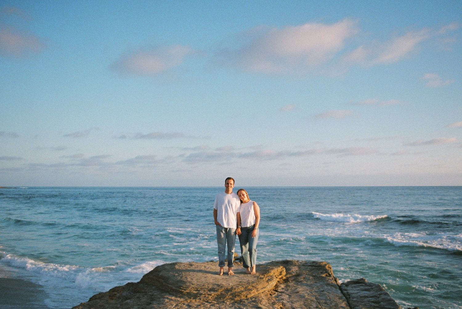 Couple standing on rocky shore with ocean waves and blue sky backdrop.