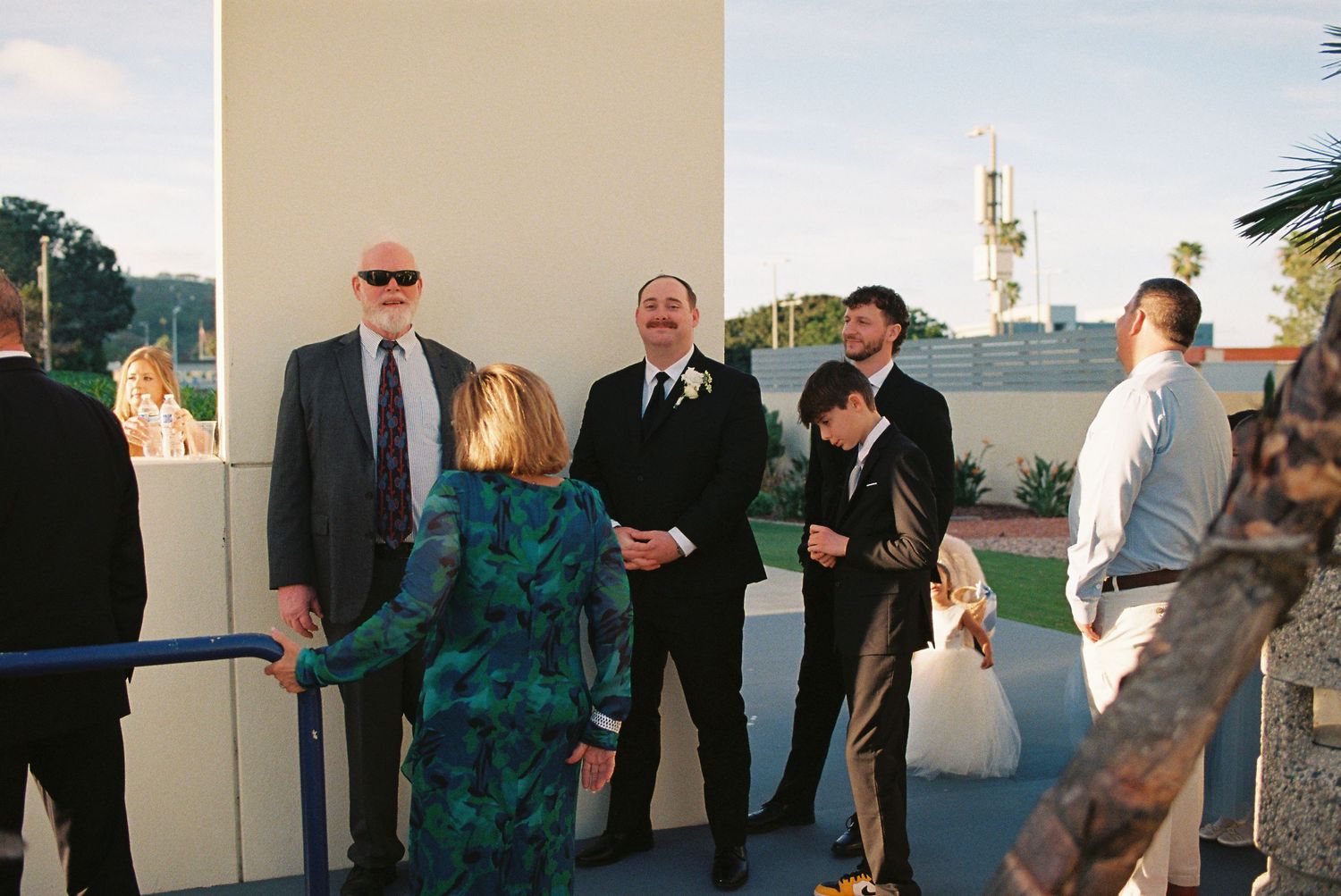 Wedding guests gathered outside venue chatting before ceremony, men in formal suits with boutonniere at coastal location.