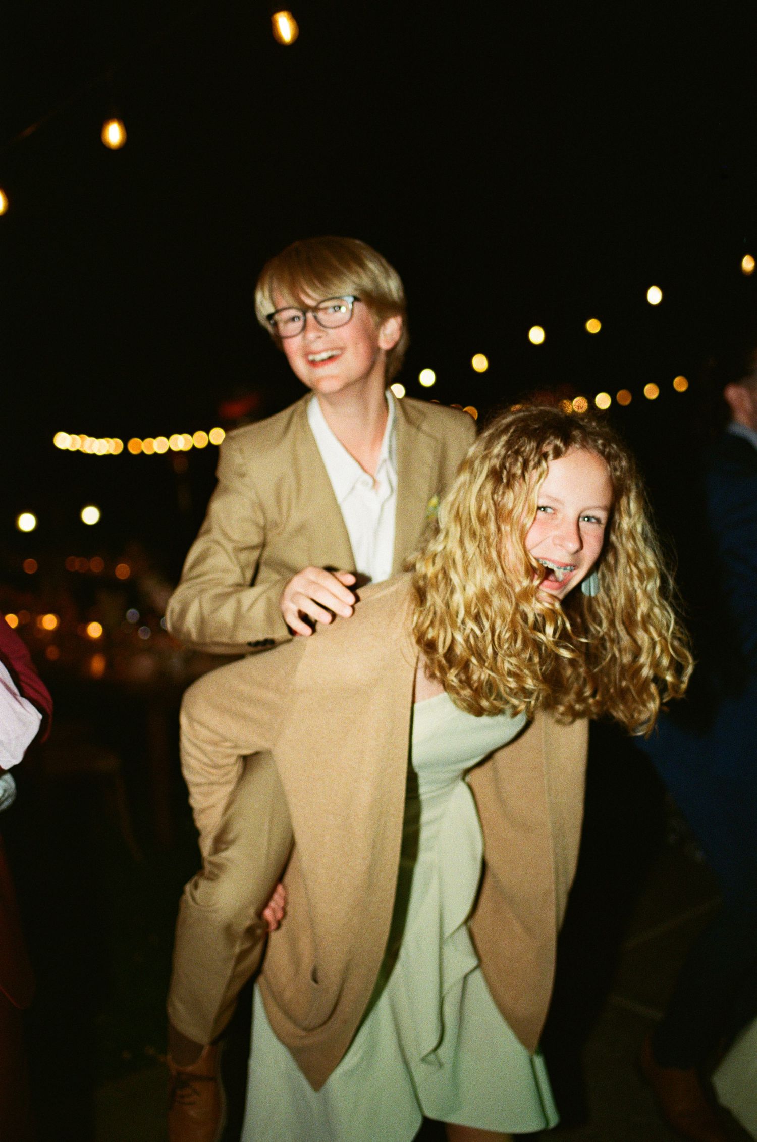 Two young wedding guests laughing as one gives the other a piggyback ride at a night reception.
