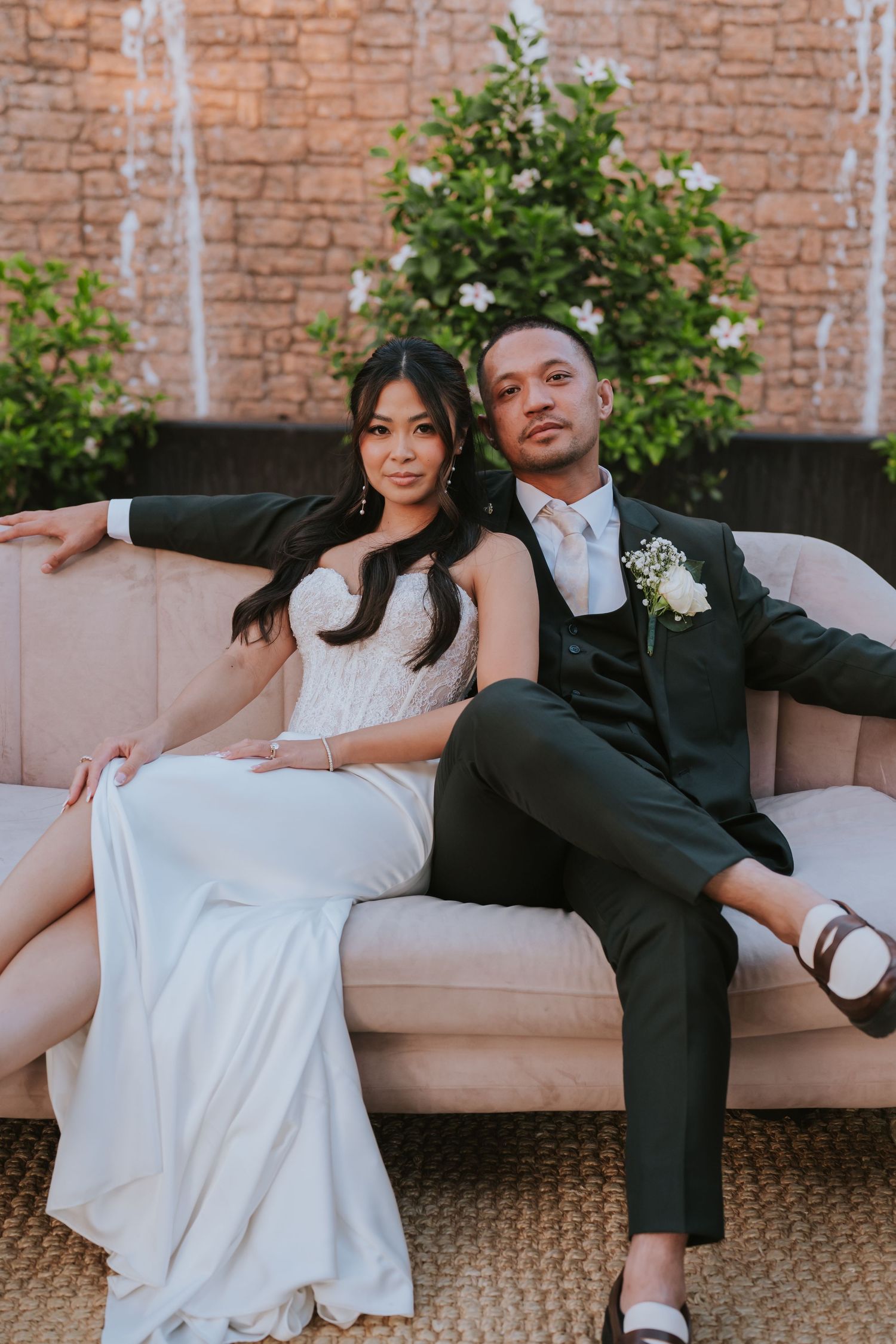 A couple in formal wedding attire poses playfully on a beige couch against a brick wall backdrop.