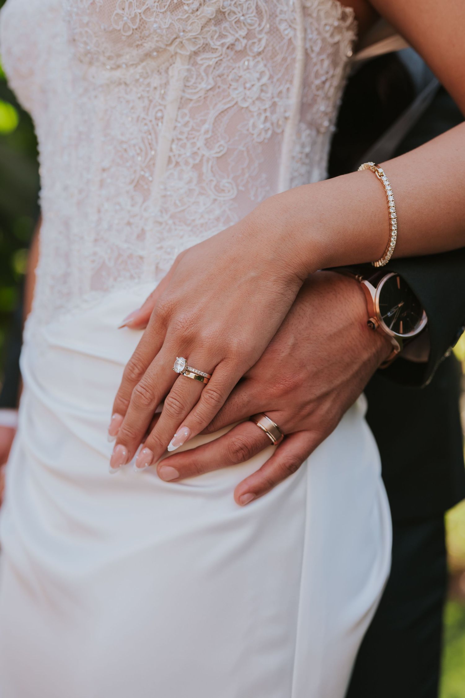Close-up photos of a bride's hands wearing a delicate diamond engagement ring and bracelet against a white lace dress.