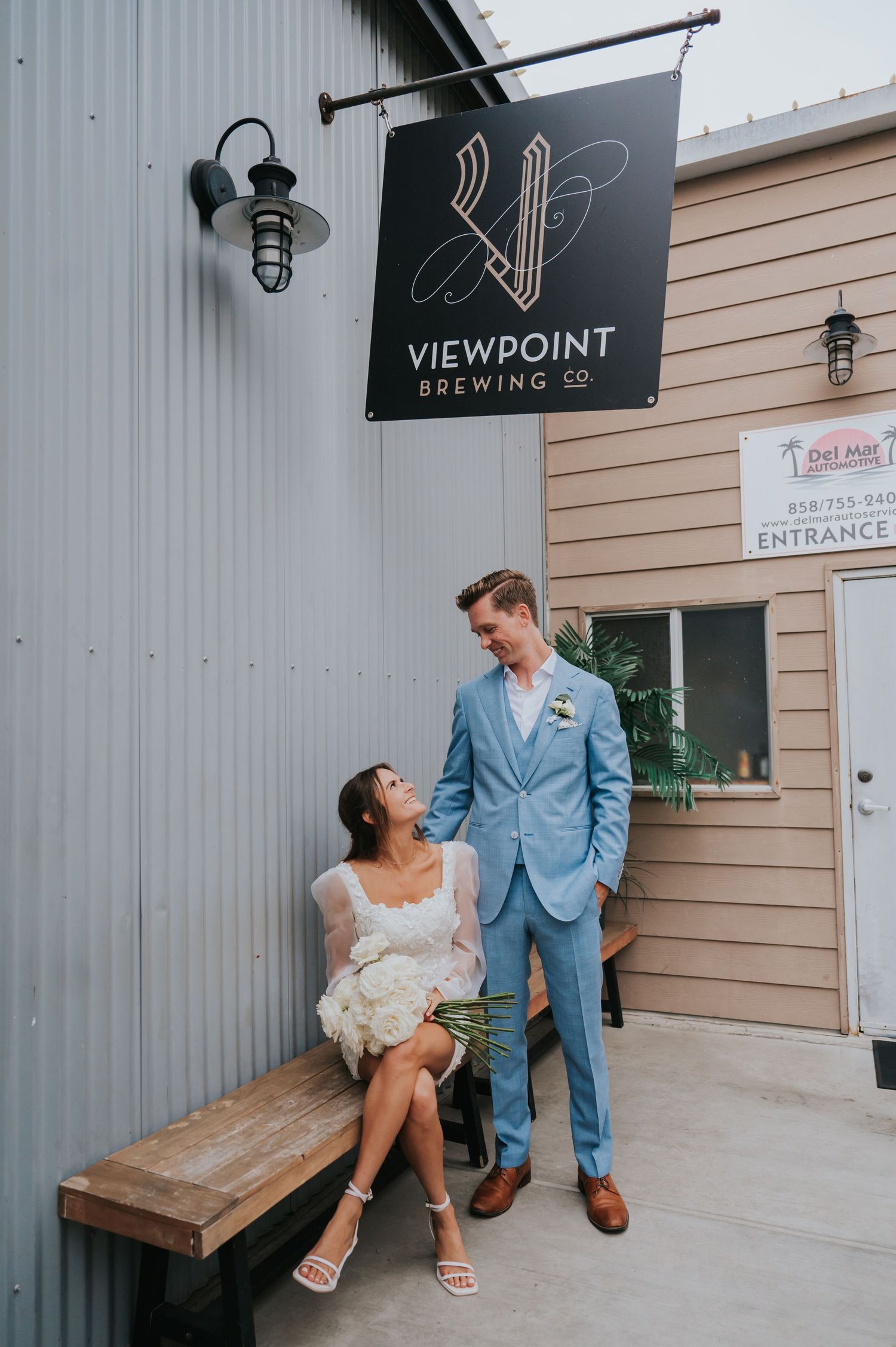 A couple poses together outside Viewpoint Brewing Co, with a wooden bench and industrial wall setting.