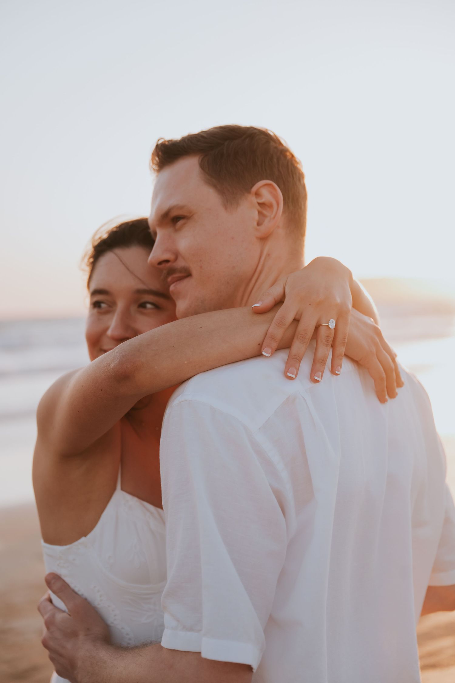 A couple embraces on a beach at sunset, the woman wearing a ring and resting her head on the man's shoulder.