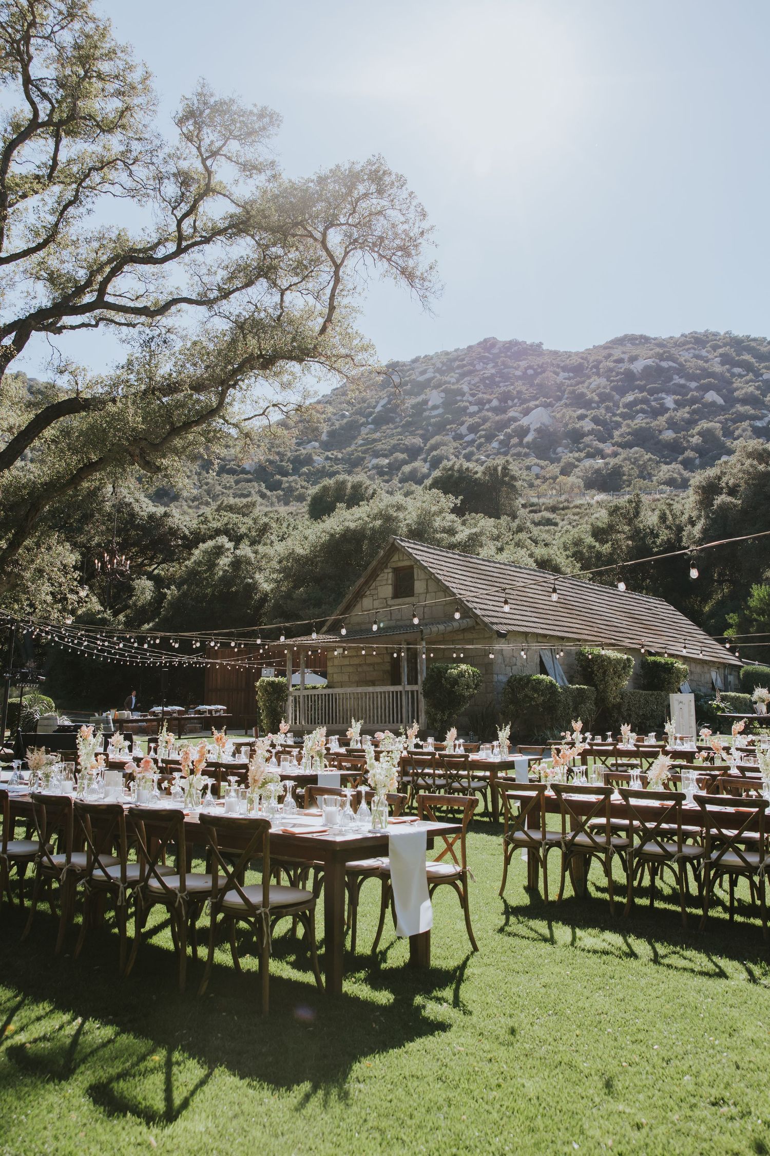 Outdoor rustic wedding reception setup on green lawn with wooden tables, string lights, barn, and mountain backdrop.