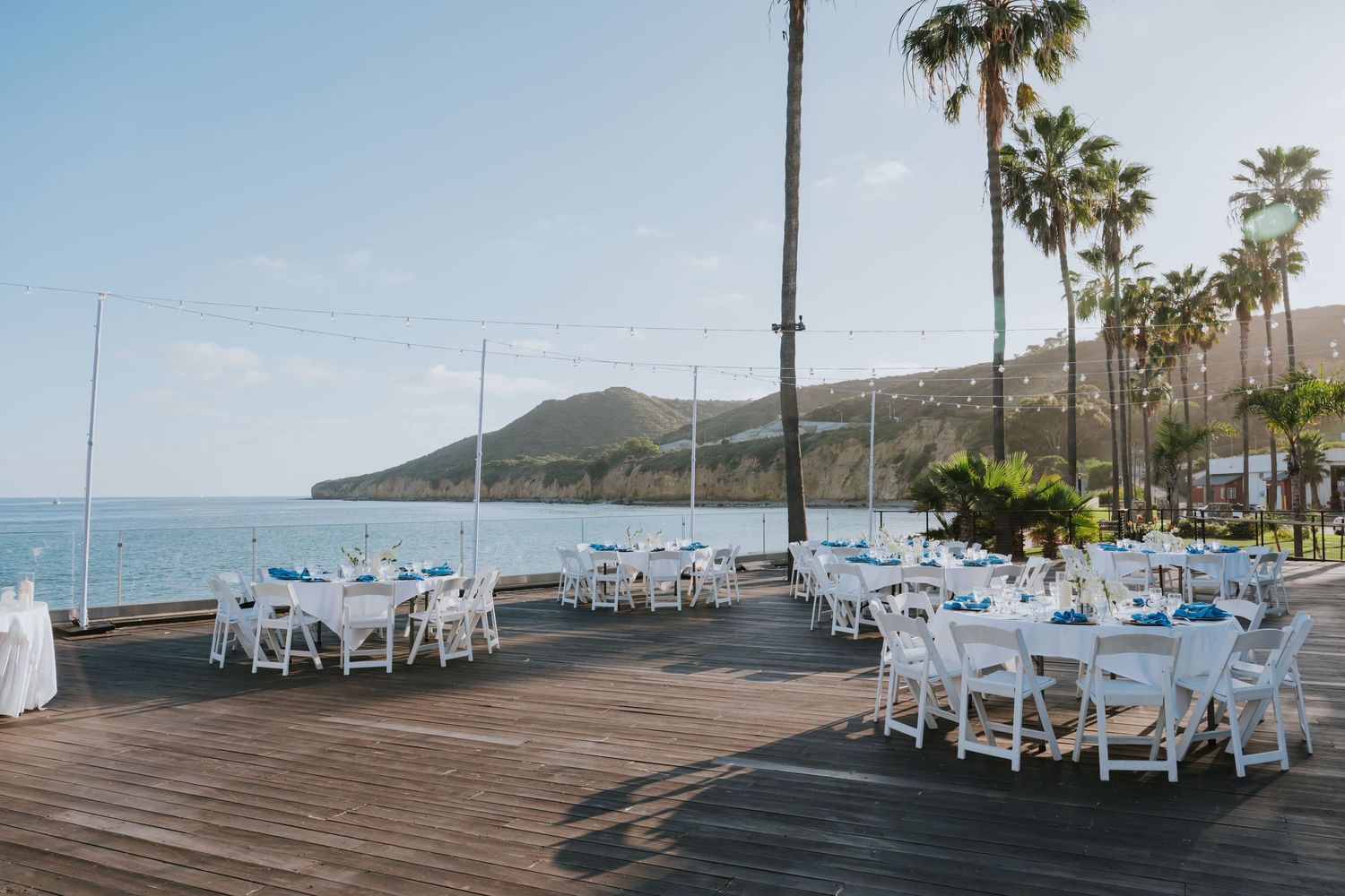 Elegant outdoor wedding reception on a wooden deck overlooking the ocean with white chairs and palm trees.