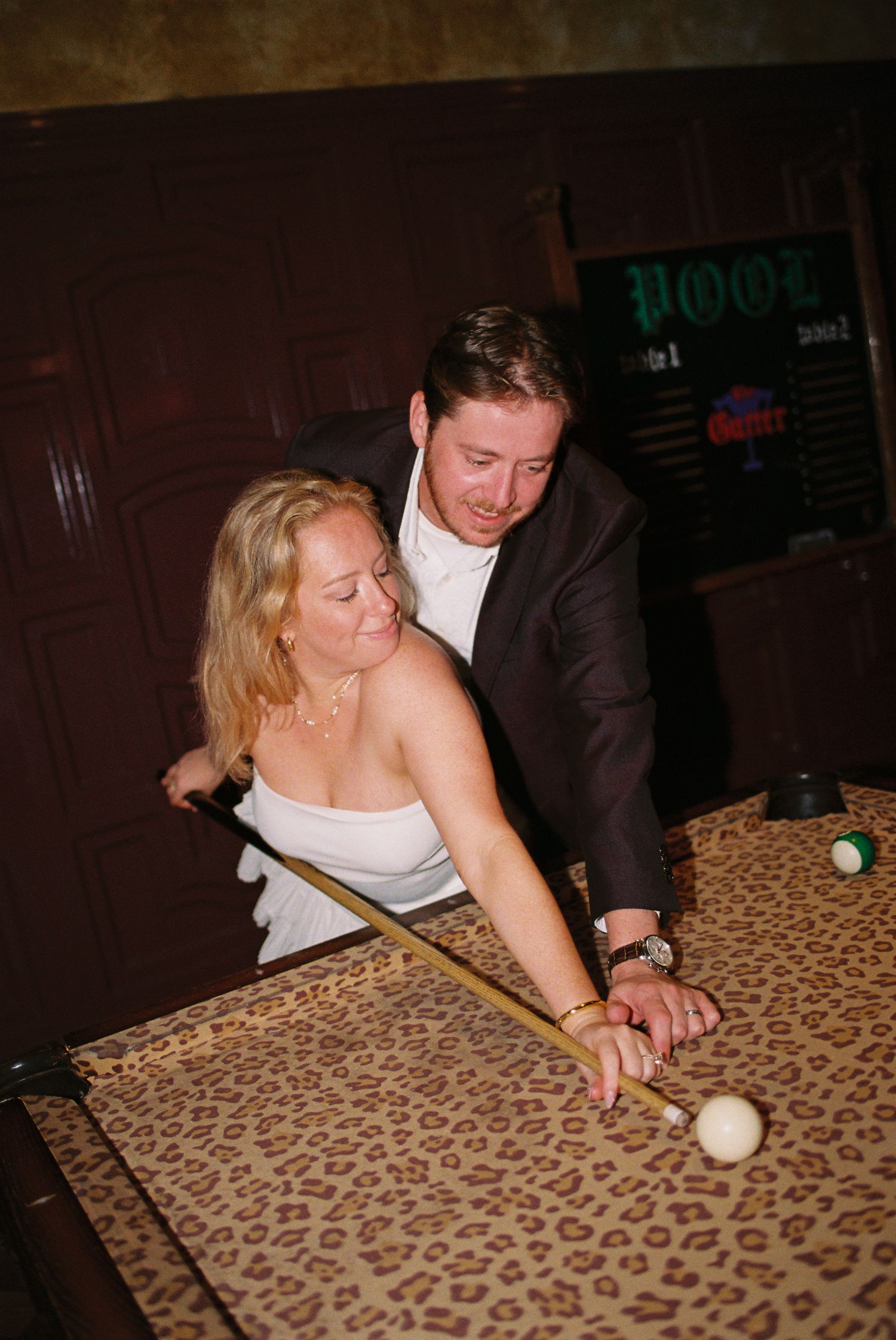 A couple playing pool together at a billiard table in a dimly lit bar.