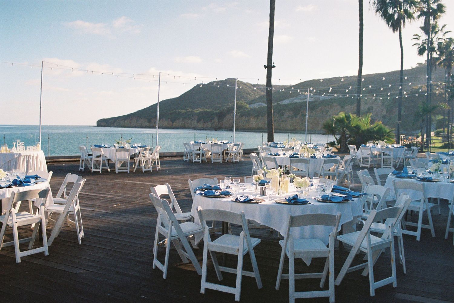 Outdoor waterfront wedding reception with white chairs and tables with blue linens on a wooden deck overlooking the ocean.
