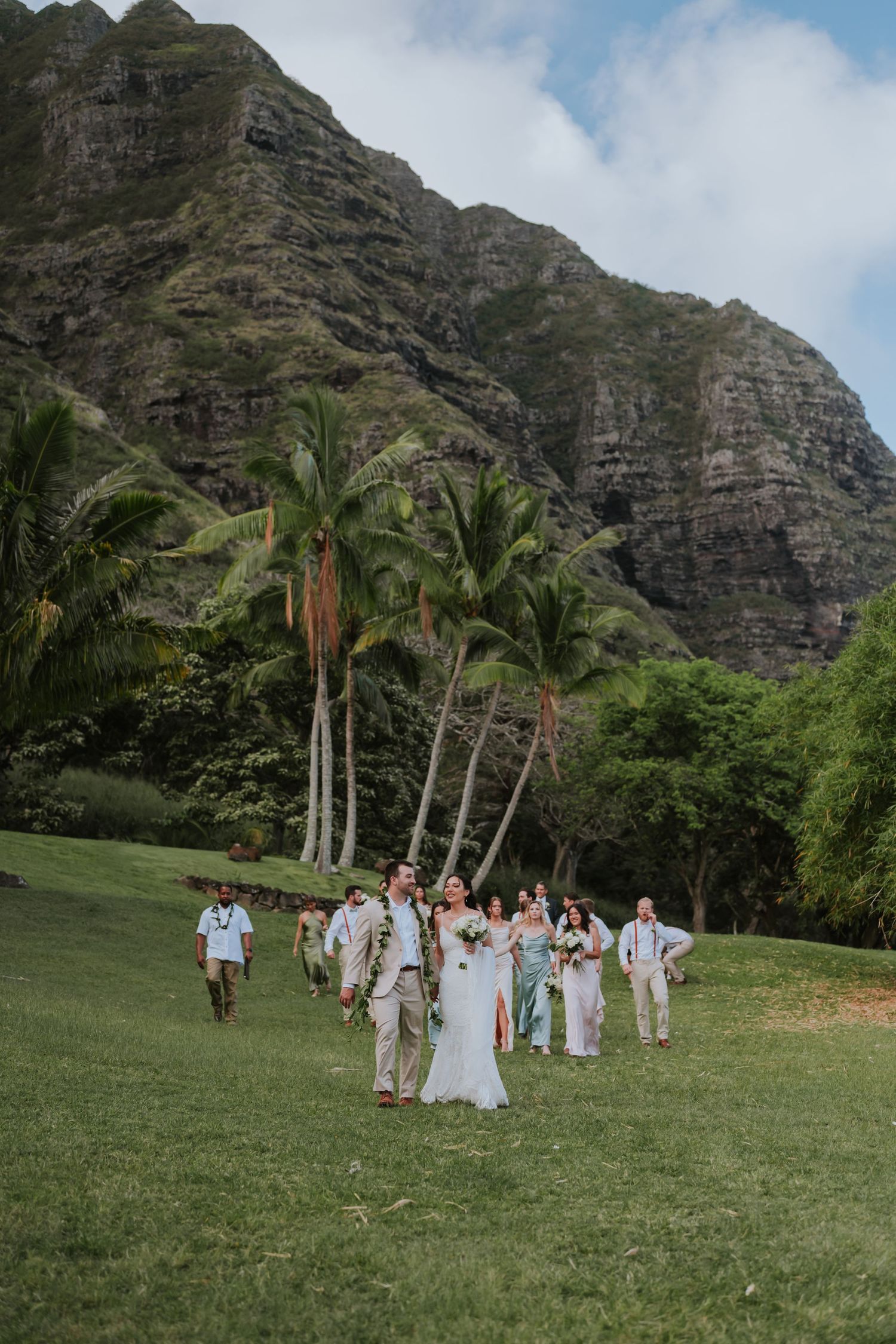 Wedding ceremony takes place on grassy lawn with dramatic mountain cliffs and palm trees in Hawaii.
