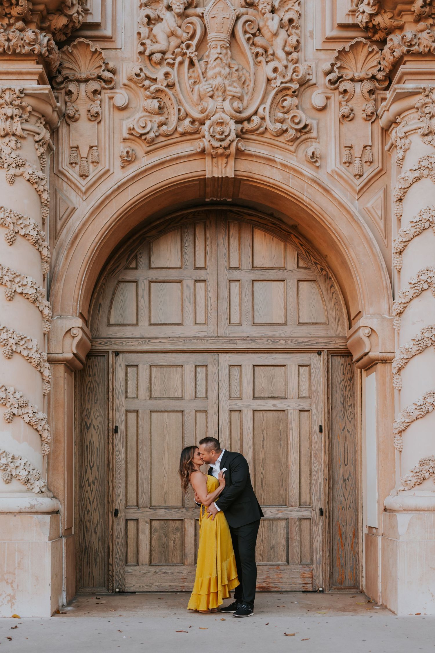 A couple embraces under an ornate baroque archway with detailed stone carvings and a large wooden door.