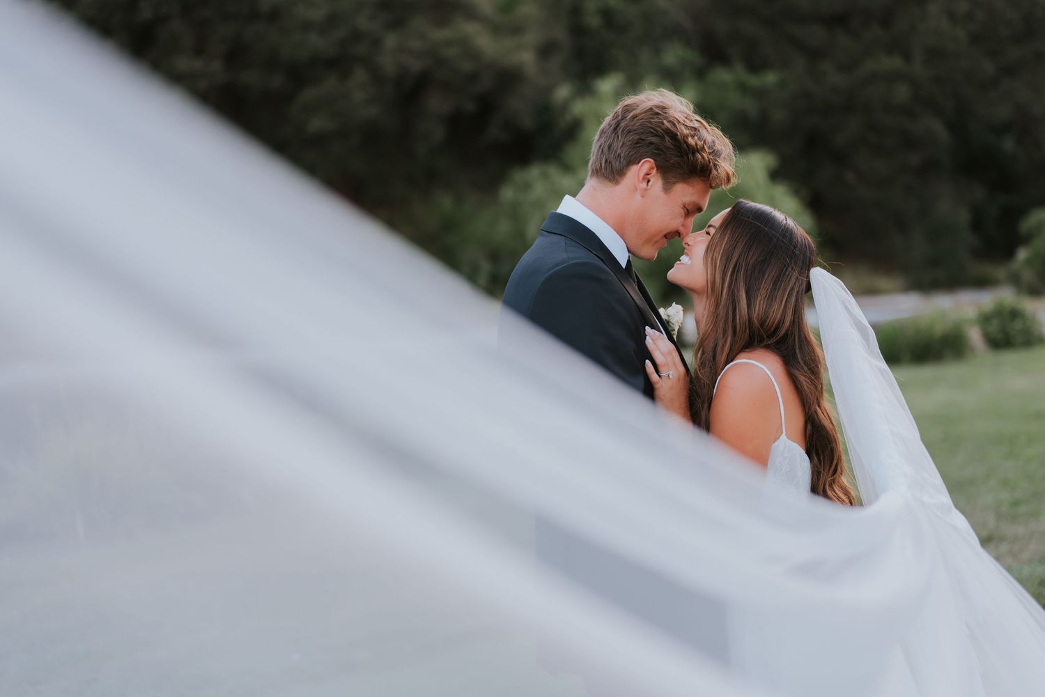 A bride and groom share an intimate moment outdoors, her veil billowing in the foreground at their wedding.