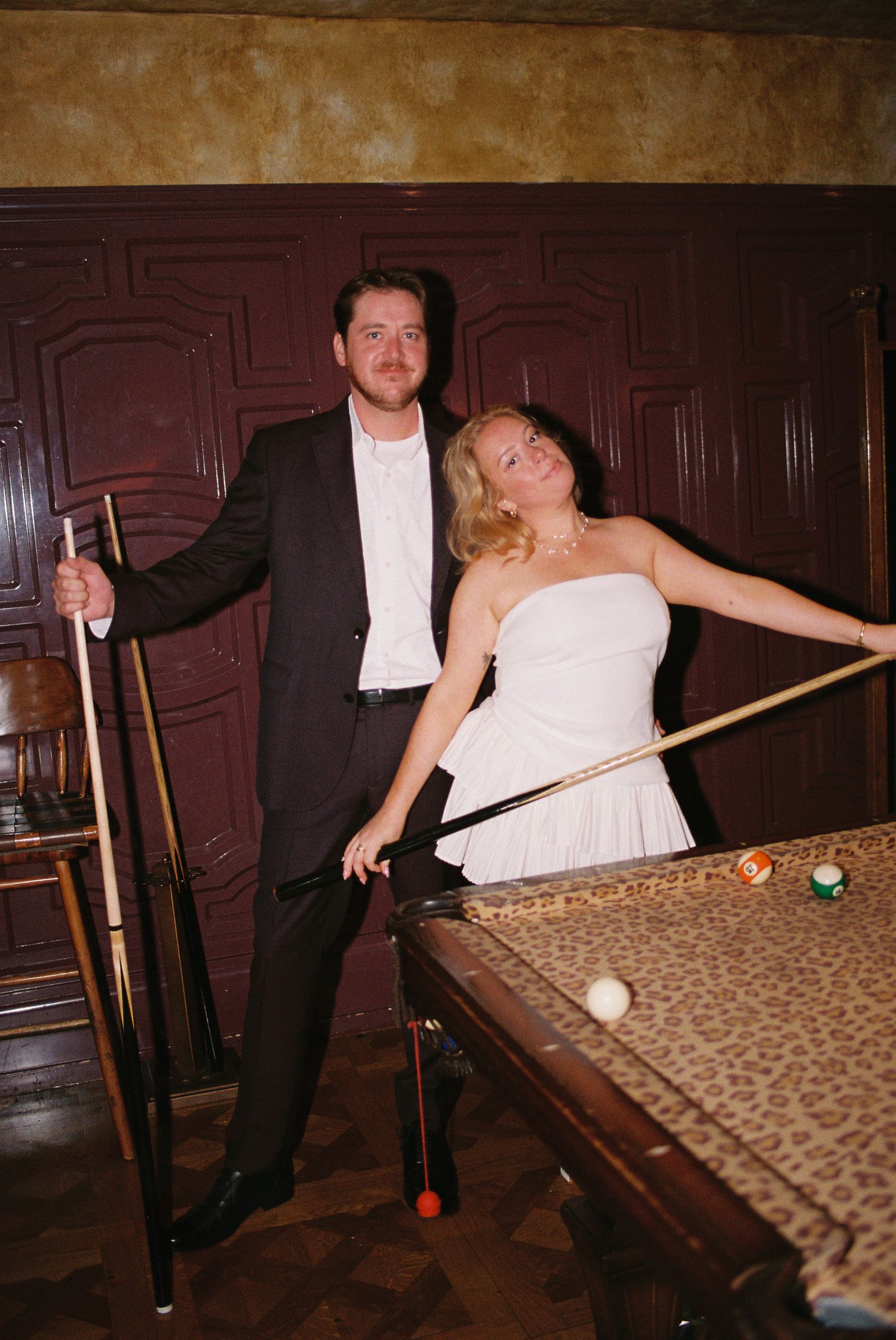 Couple playing pool in elegant billiard room, woman in white strapless dress leaning on leopard-print table.