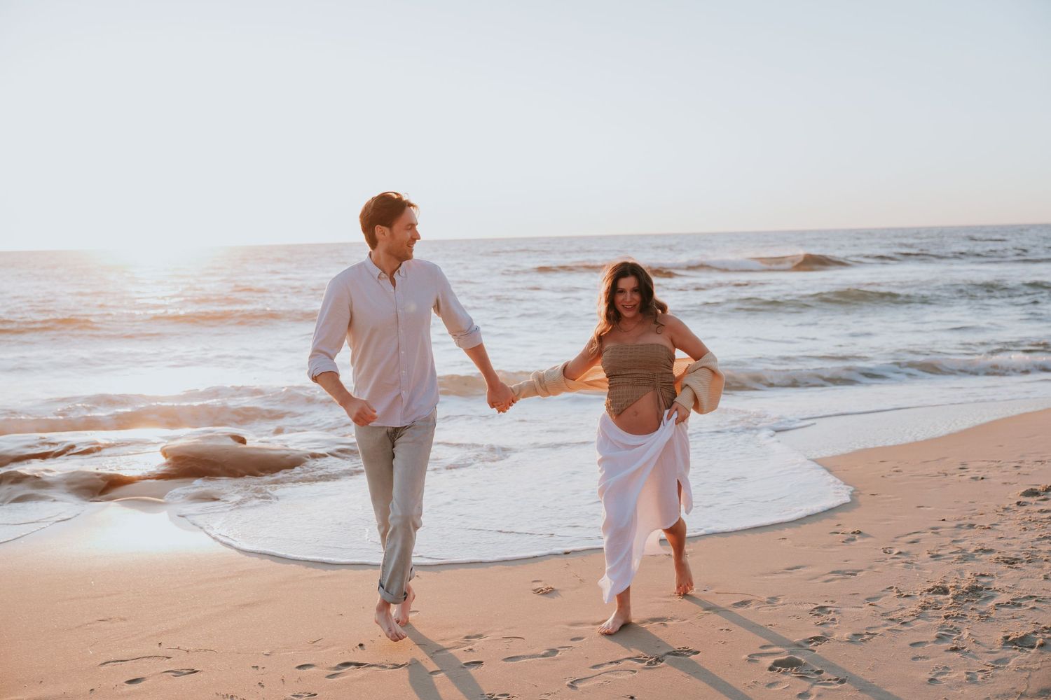 A couple in white outfits walks hand in hand along a sandy beach at sunset with ocean waves in the background.