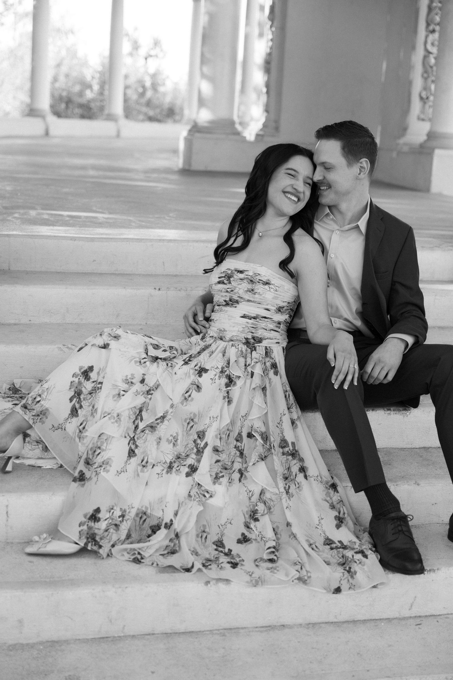Black and white photo of a couple sitting on steps, smiling and embracing, woman in floral dress.
