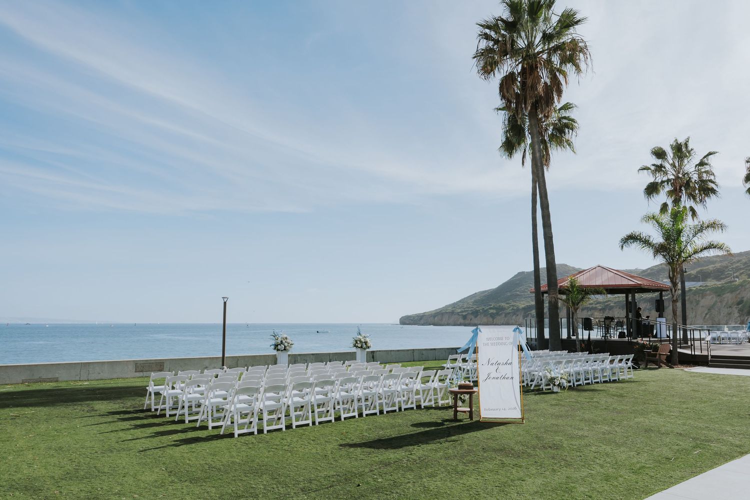 Outdoor beachside wedding ceremony setup with white chairs on green lawn, palm trees, and ocean views under a clear blue sky.
