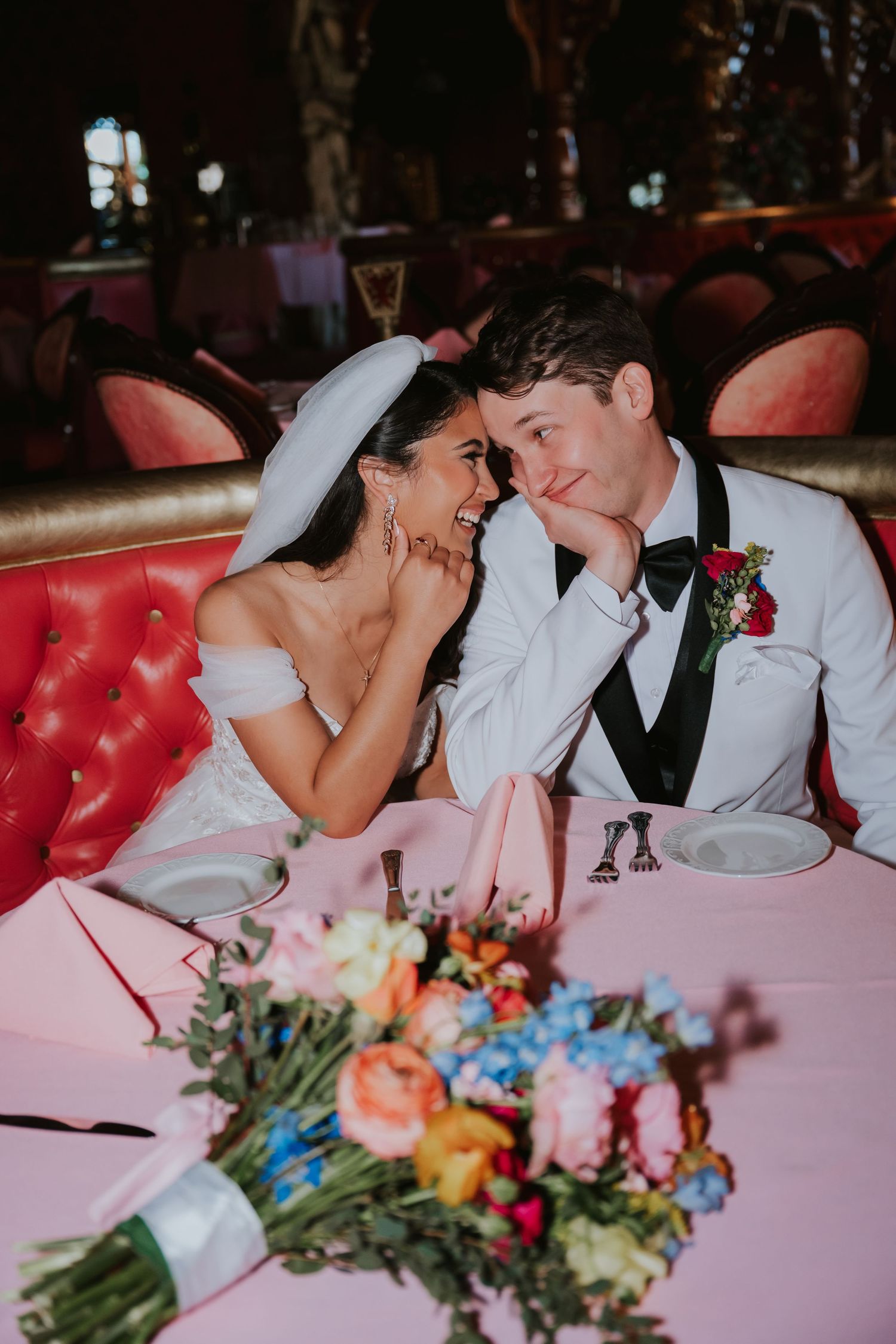 A romantic moment shared between newlyweds on a pink velvet couch with colorful floral arrangements in the foreground.