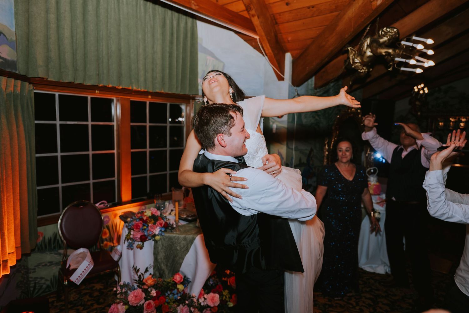 Wedding guests dance and celebrate on a dimly lit dance floor at a reception venue.