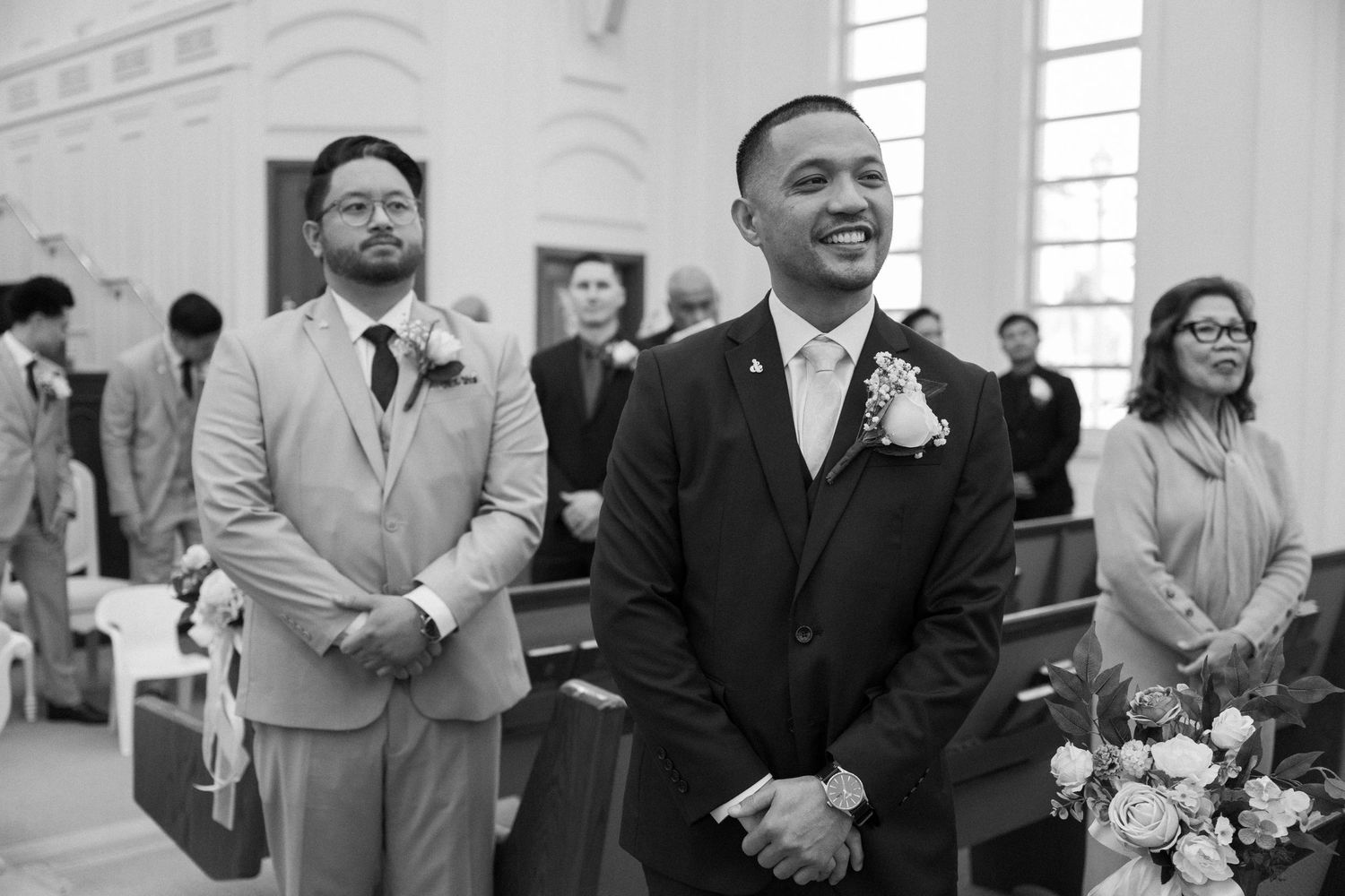 A group of formally dressed people stand together during a wedding ceremony in a church with stained glass windows.