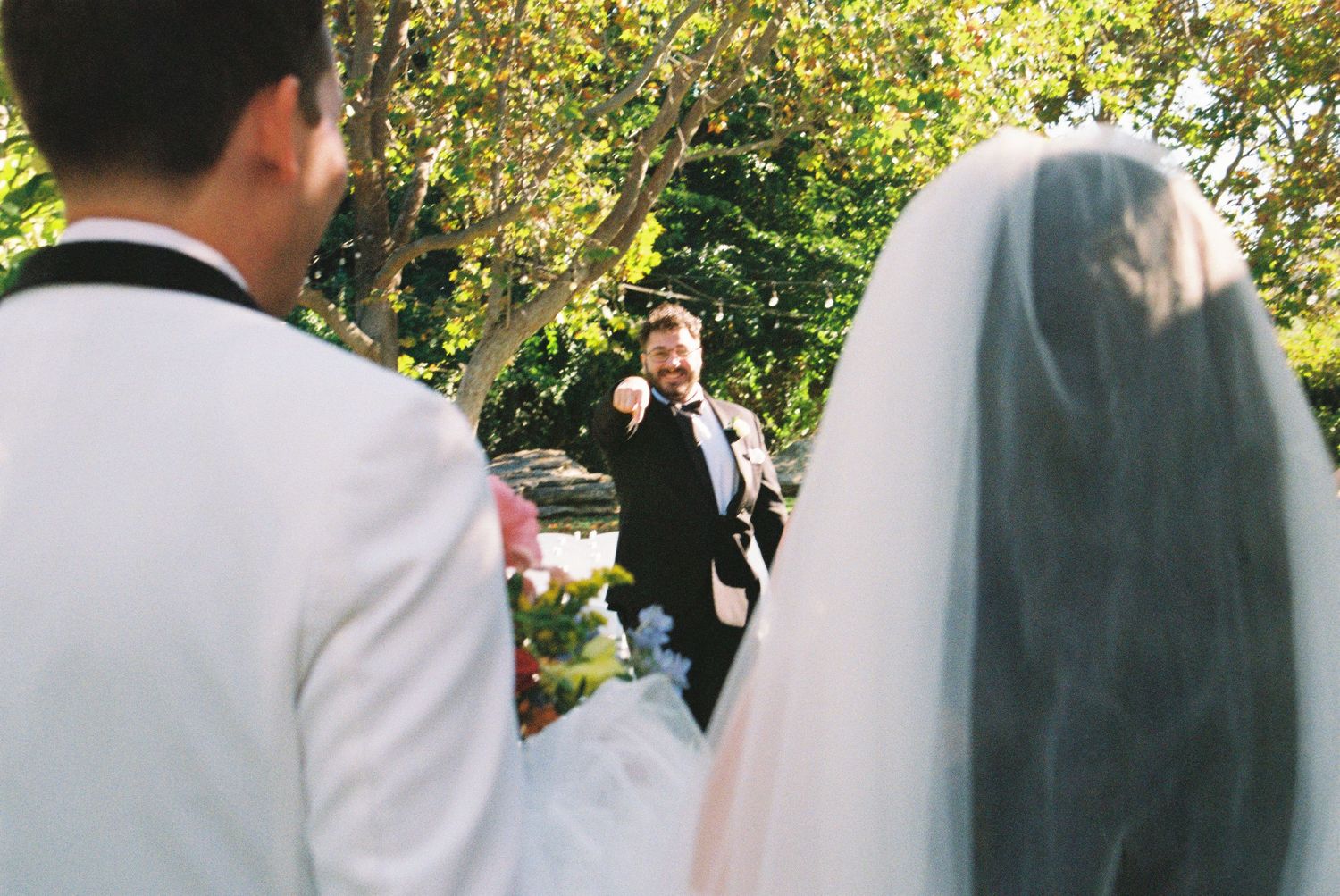 Outdoor wedding ceremony with bride in veil and groom in white suit facing officiant under green trees.