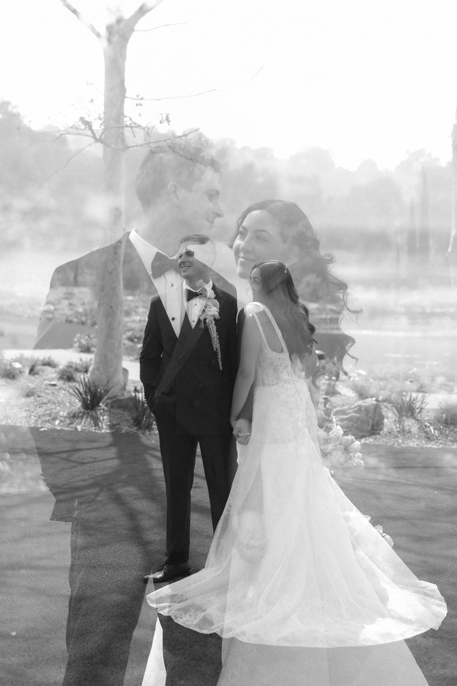 Black and white double exposure wedding photo of bride in gown and groom in tuxedo smiling at each other outdoors.