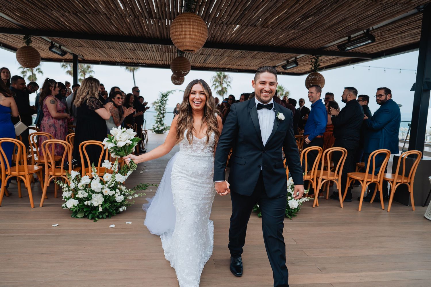 Wedding ceremony procession in an oceanfront covered venue with palm trees in background.