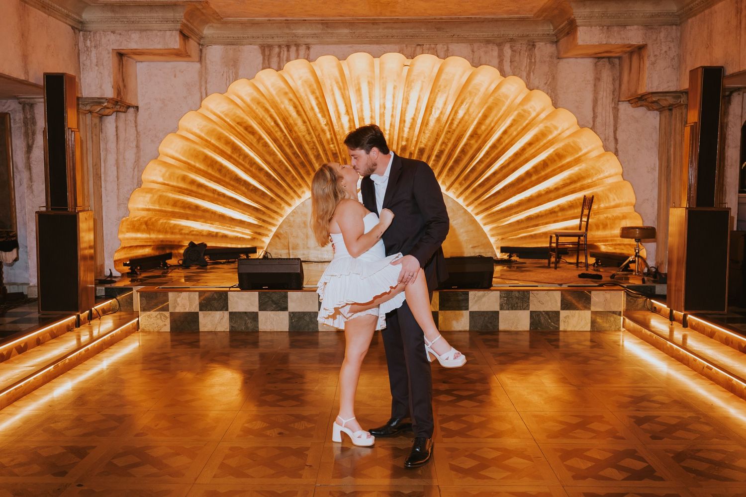 Bride and groom share a romantic dip dance on a golden-lit Art Deco dance floor with ornate shell backdrop.