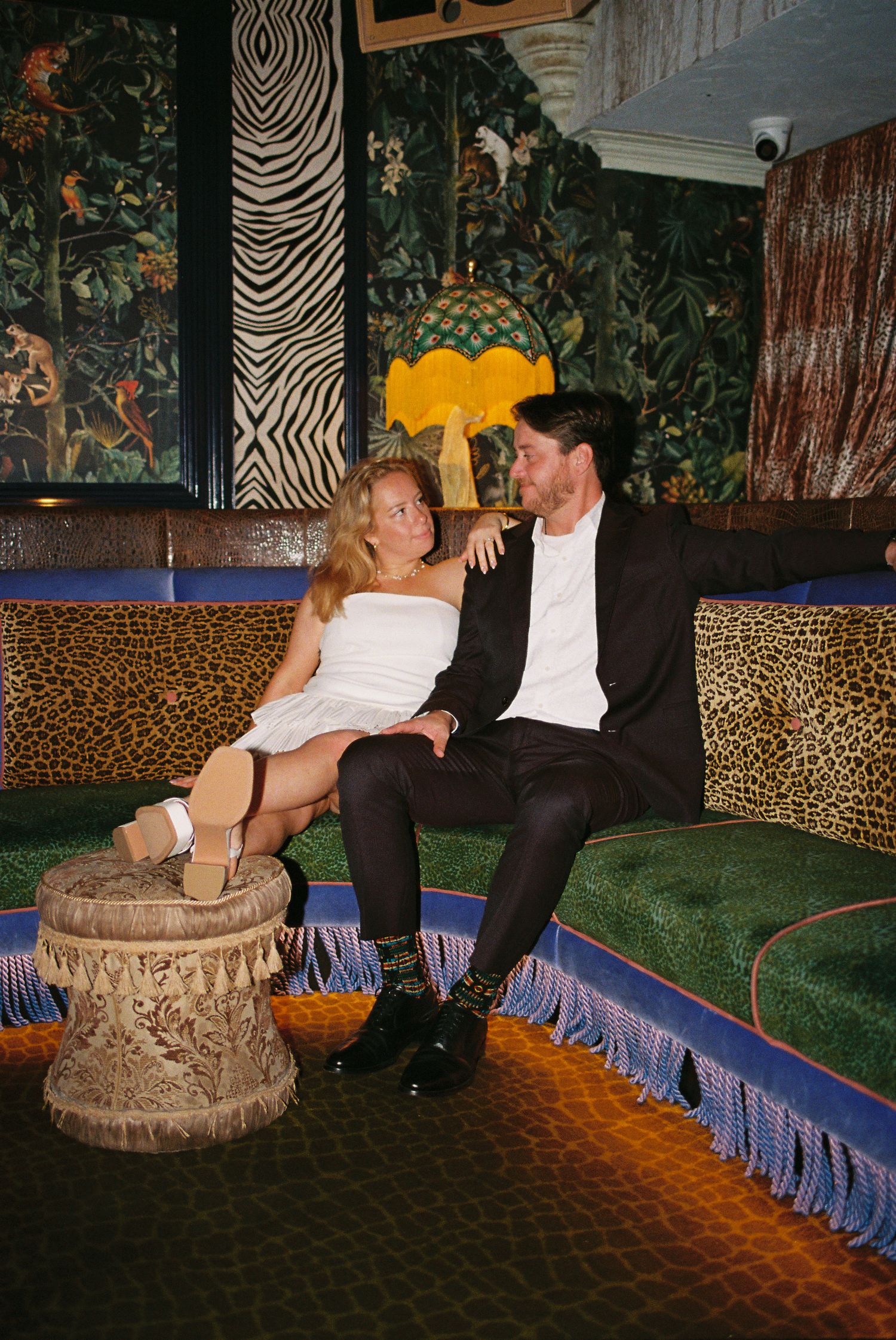 A couple sits intimately on a green velvet sofa in a vibrant, eclectic lounge with zebra and leopard print decor.