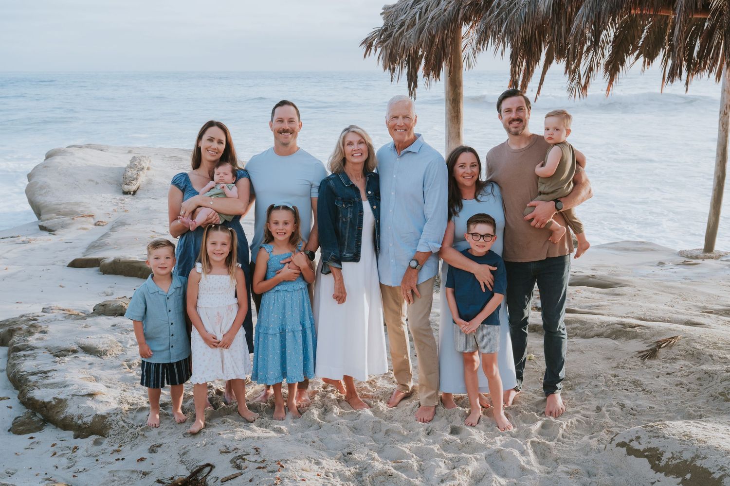 Large family group portrait on a sandy beach near rocky shores, posing together for a casual vacation photo.