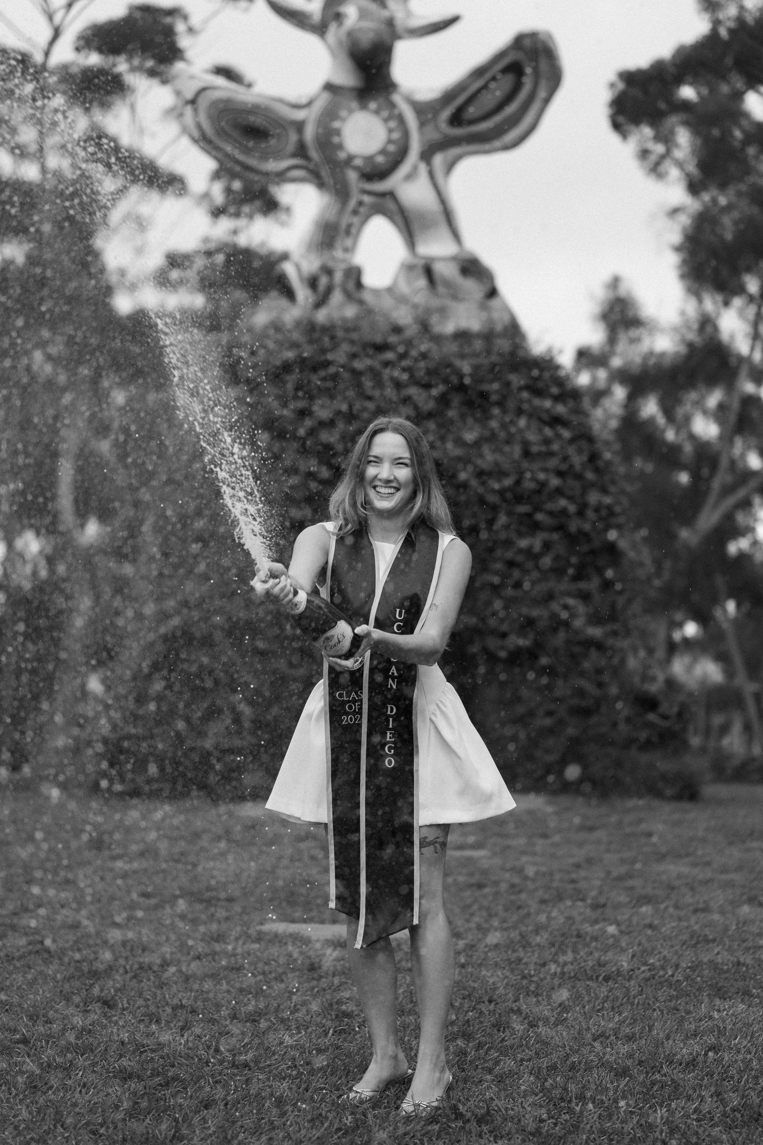 Black and white photo of a smiling graduate in white dress and sash popping champagne outdoors.