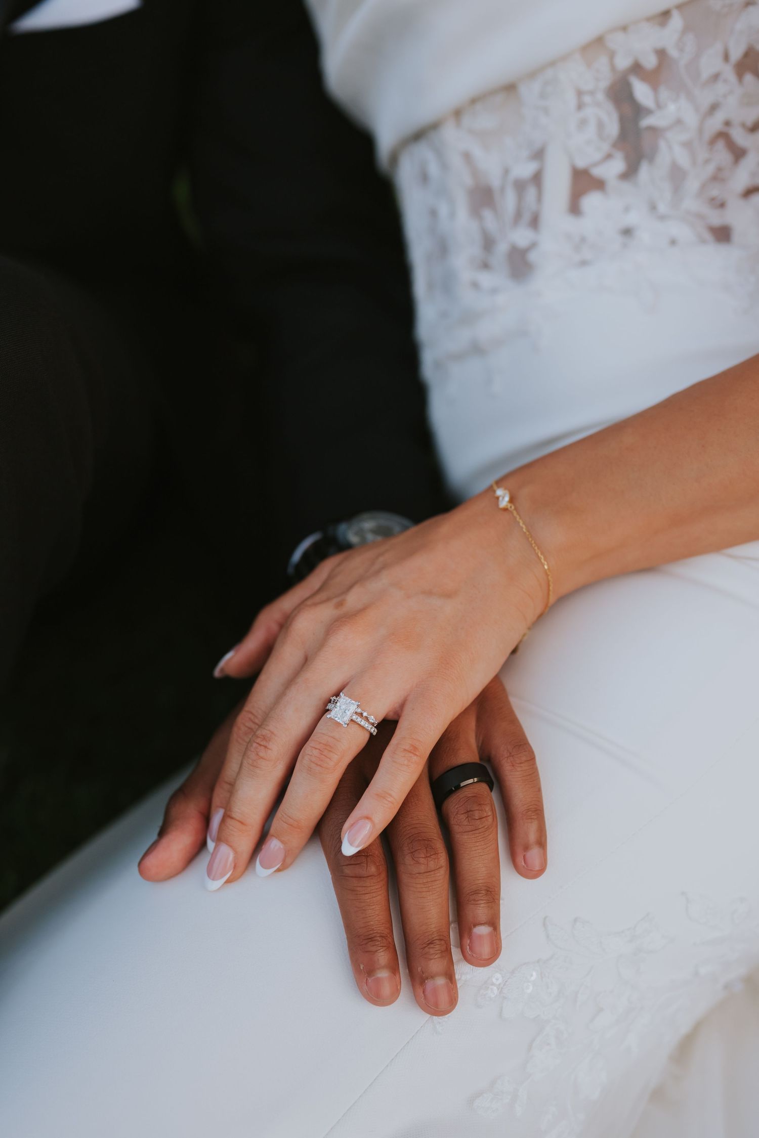 Close-up of bride and groom hands showing elegant diamond engagement ring and black wedding band on wedding day.