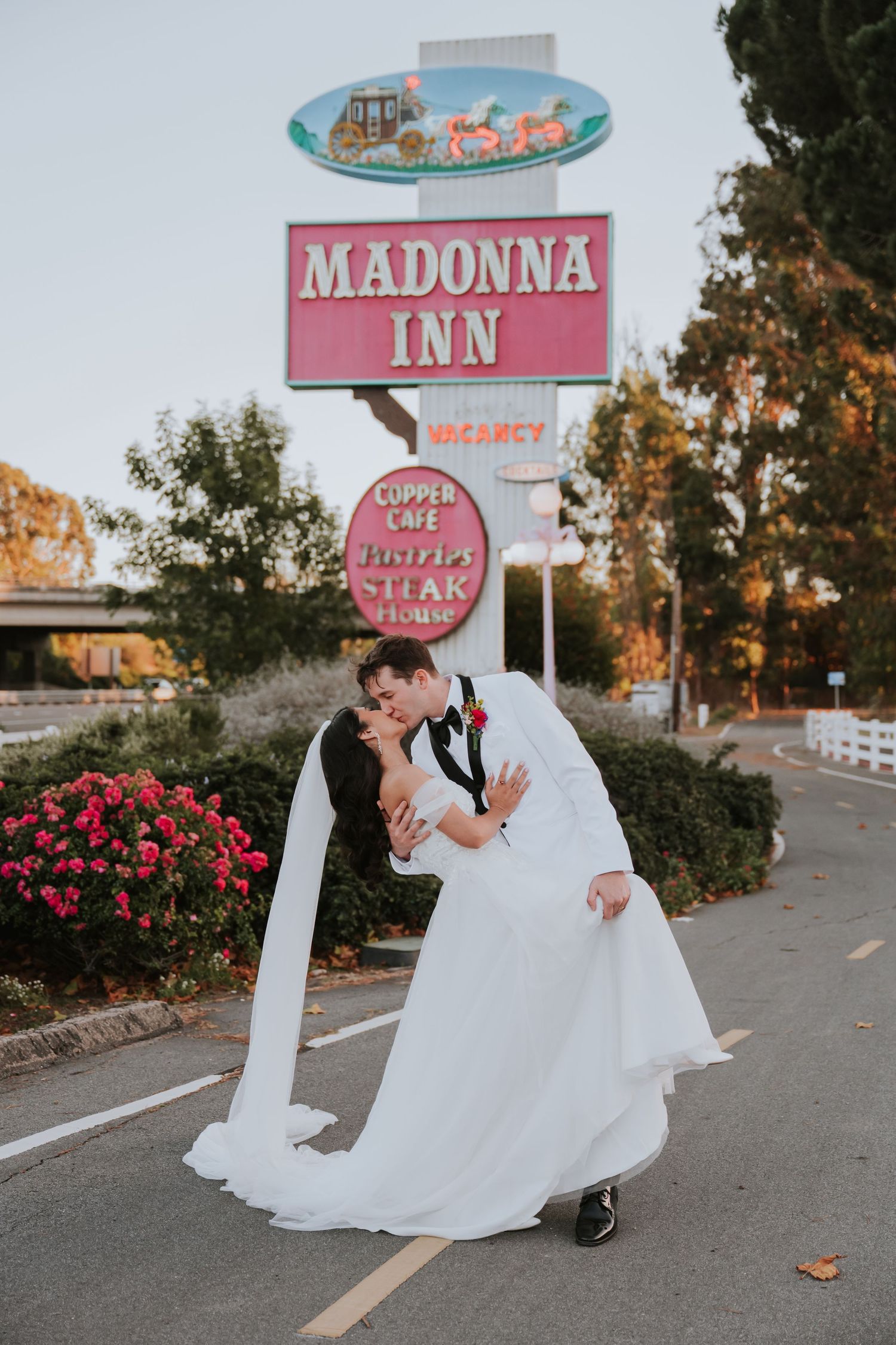 Romantic wedding couple poses in front of vintage Madonna Inn sign with pink roses in foreground at sunset.