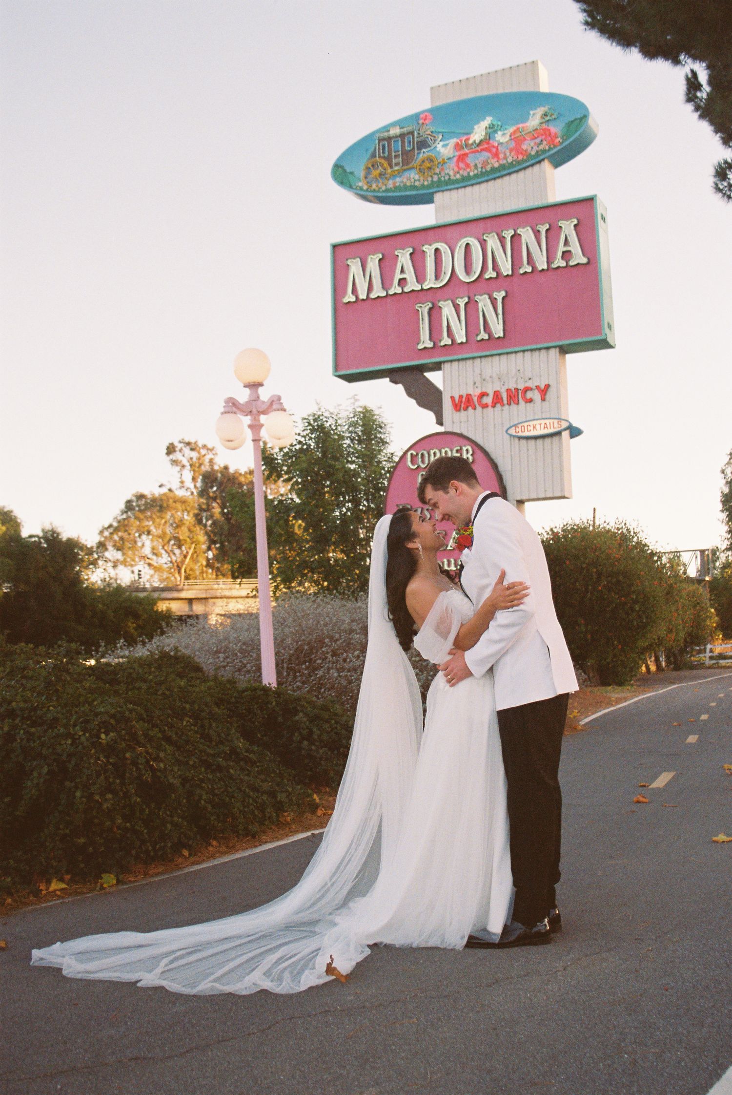Newlywed couple kissing in wedding attire in front of the iconic Madonna Inn sign in San Luis Obispo, CA.