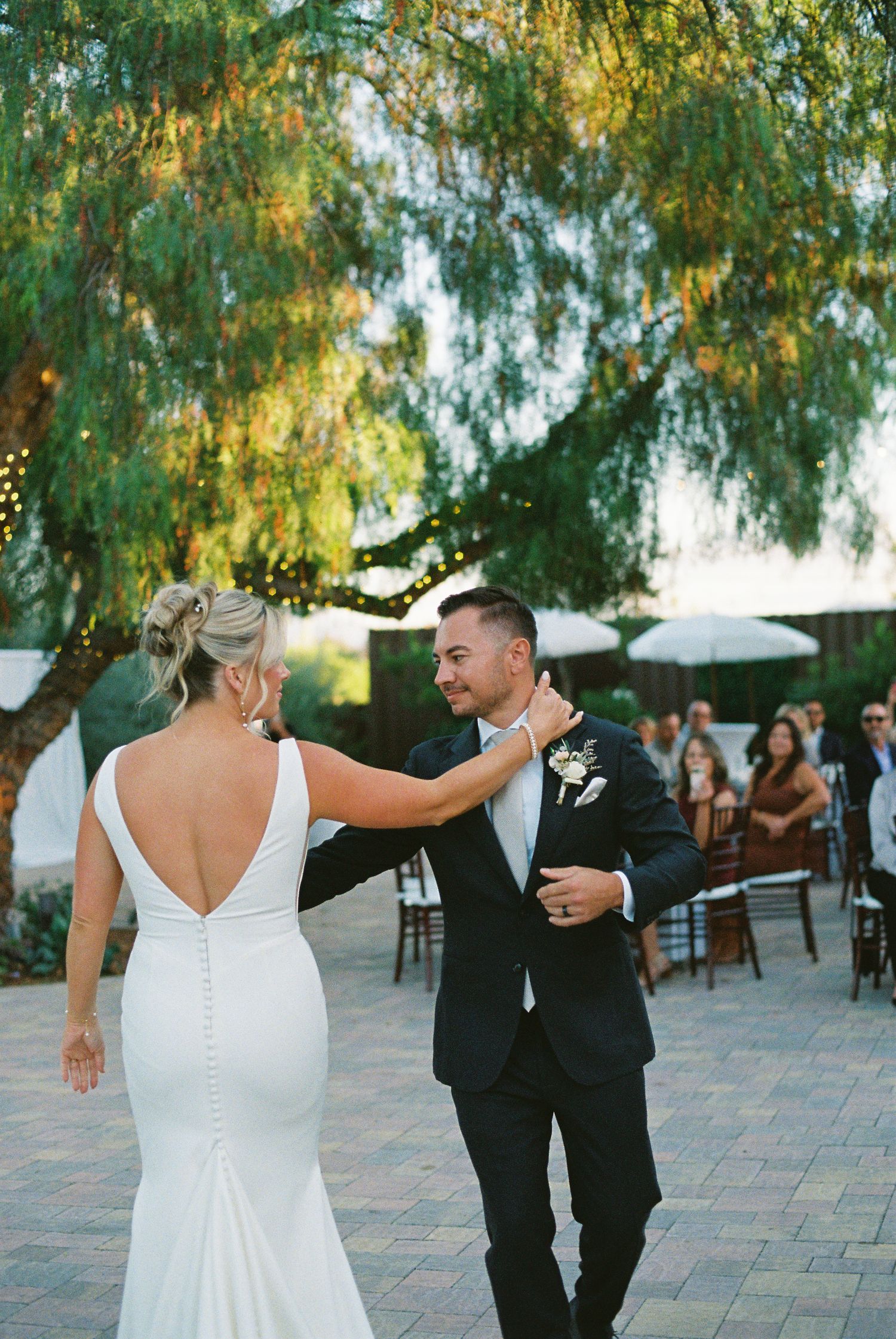 Bride and groom share their first dance outdoors under lush green trees at an elegant wedding reception.