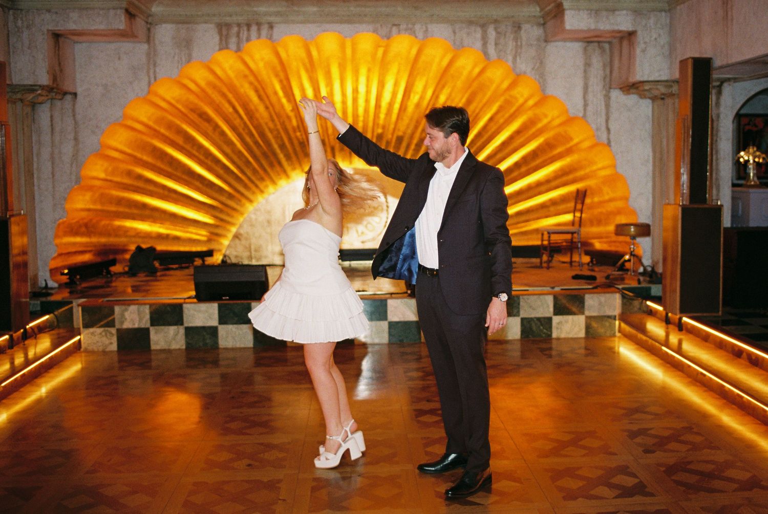 Couple dancing elegantly in a ballroom with a stunning golden fan-shaped backdrop and warm ambient lighting.