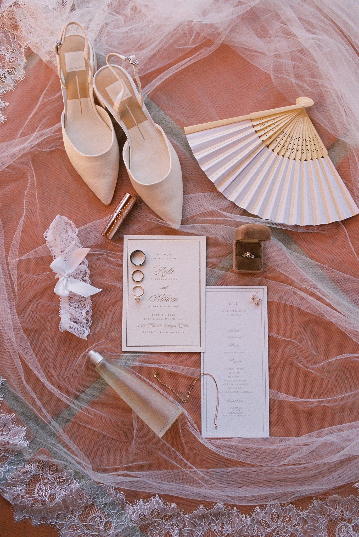 Elegant wedding flat lay with bridal shoes, fan, rings, invitation suite, garter, and veil on pink backdrop.