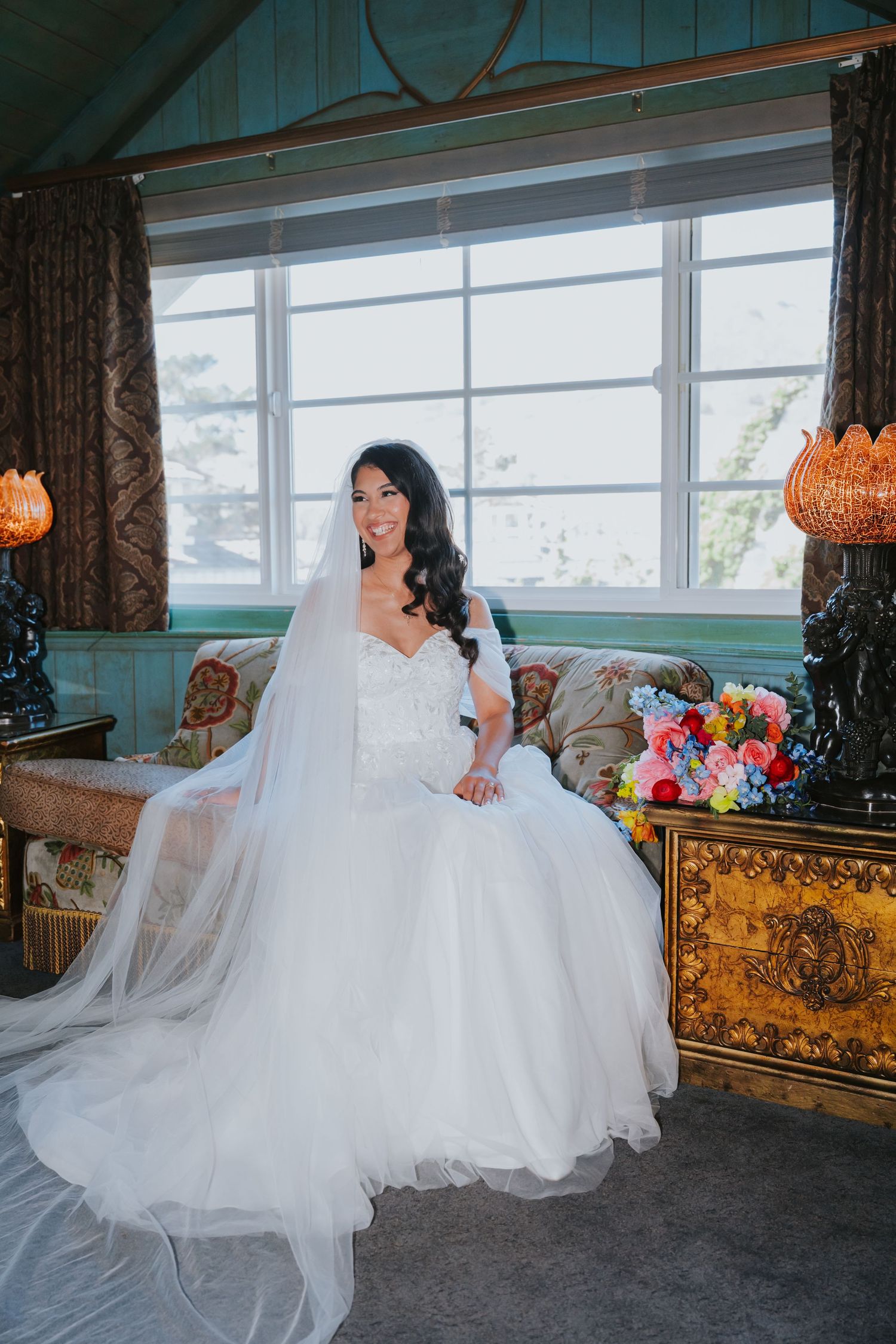 A bride in a flowing white wedding gown sits elegantly on a vintage couch near large windows.