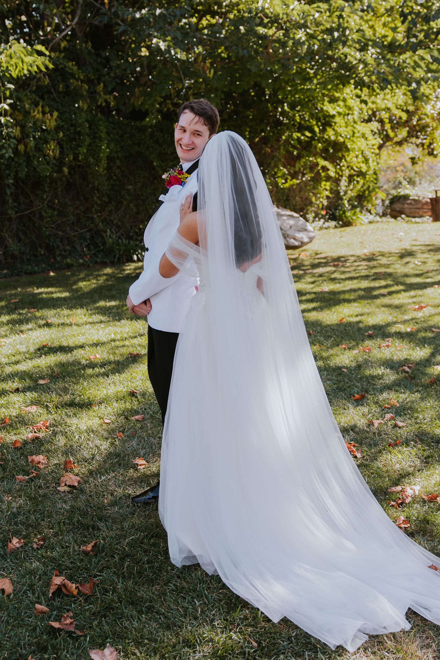 Couple in wedding attire share intimate moment on grassy lawn surrounded by autumn trees.