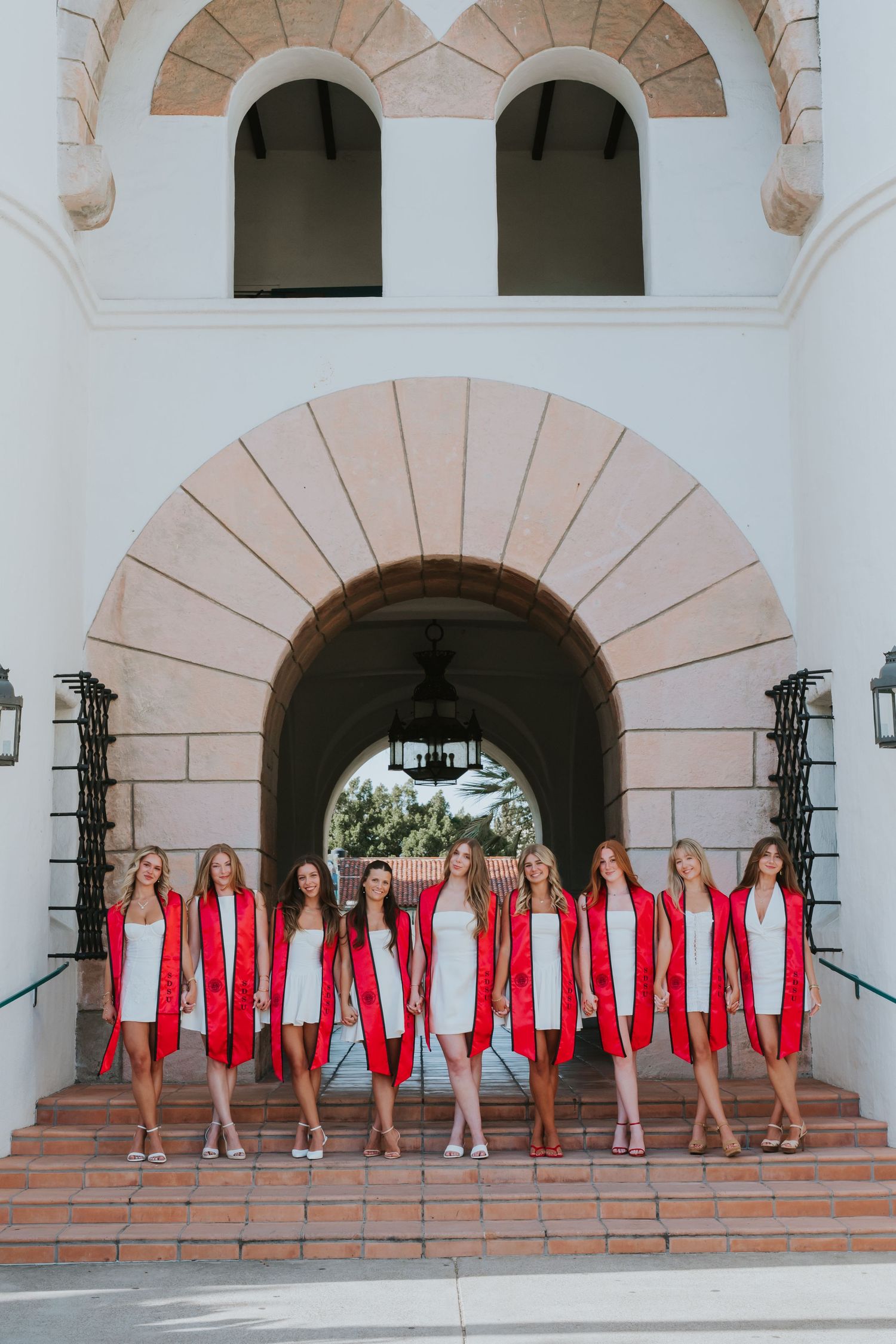 Group of women in white dresses and red graduation stoles posing on steps of an arched building.