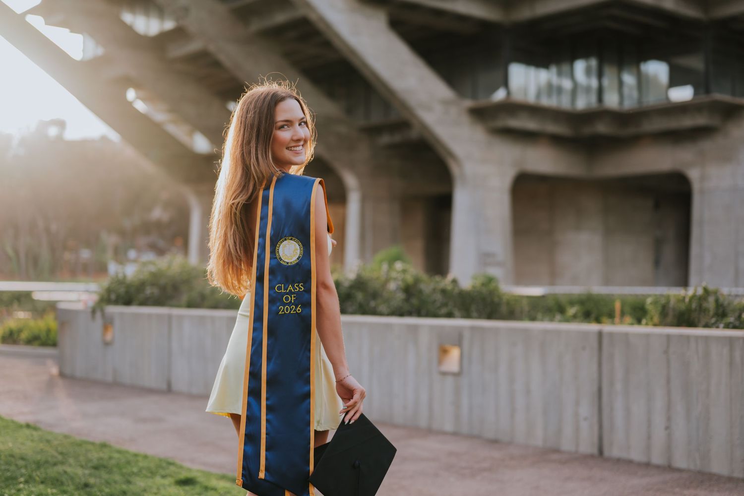 Smiling female graduate in blue and gold regalia poses outdoors at sunny campus ceremony.