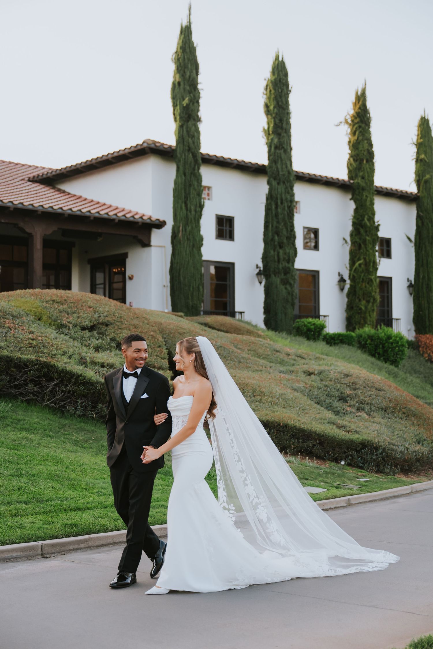 Bride and groom walking hand in hand outside elegant white Spanish-style wedding venue with tall cypress trees.