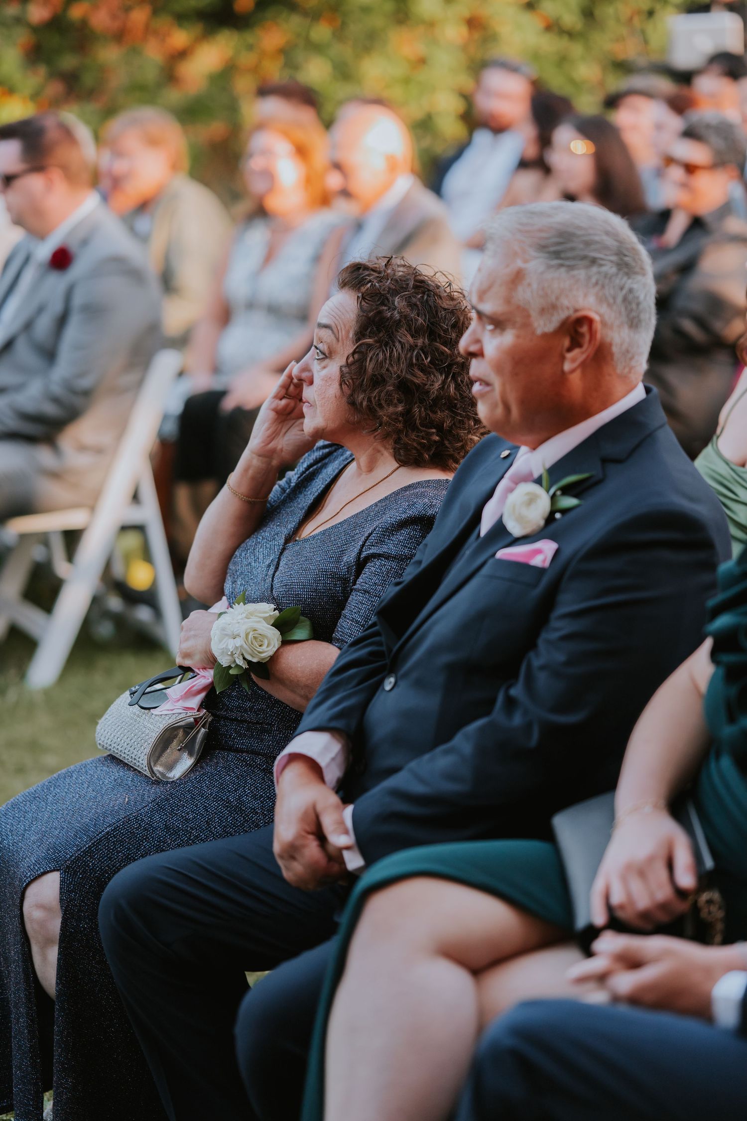 Guests seated in white chairs watch an emotional outdoor wedding ceremony at sunset with autumn foliage in background.