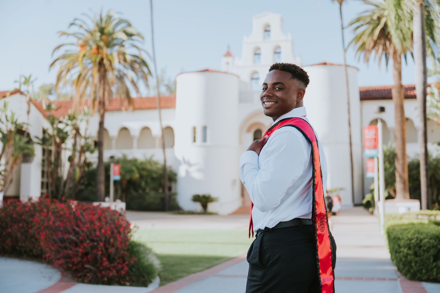 Young Black man in graduation attire smiles outdoors near a Spanish-style university building with palm trees.