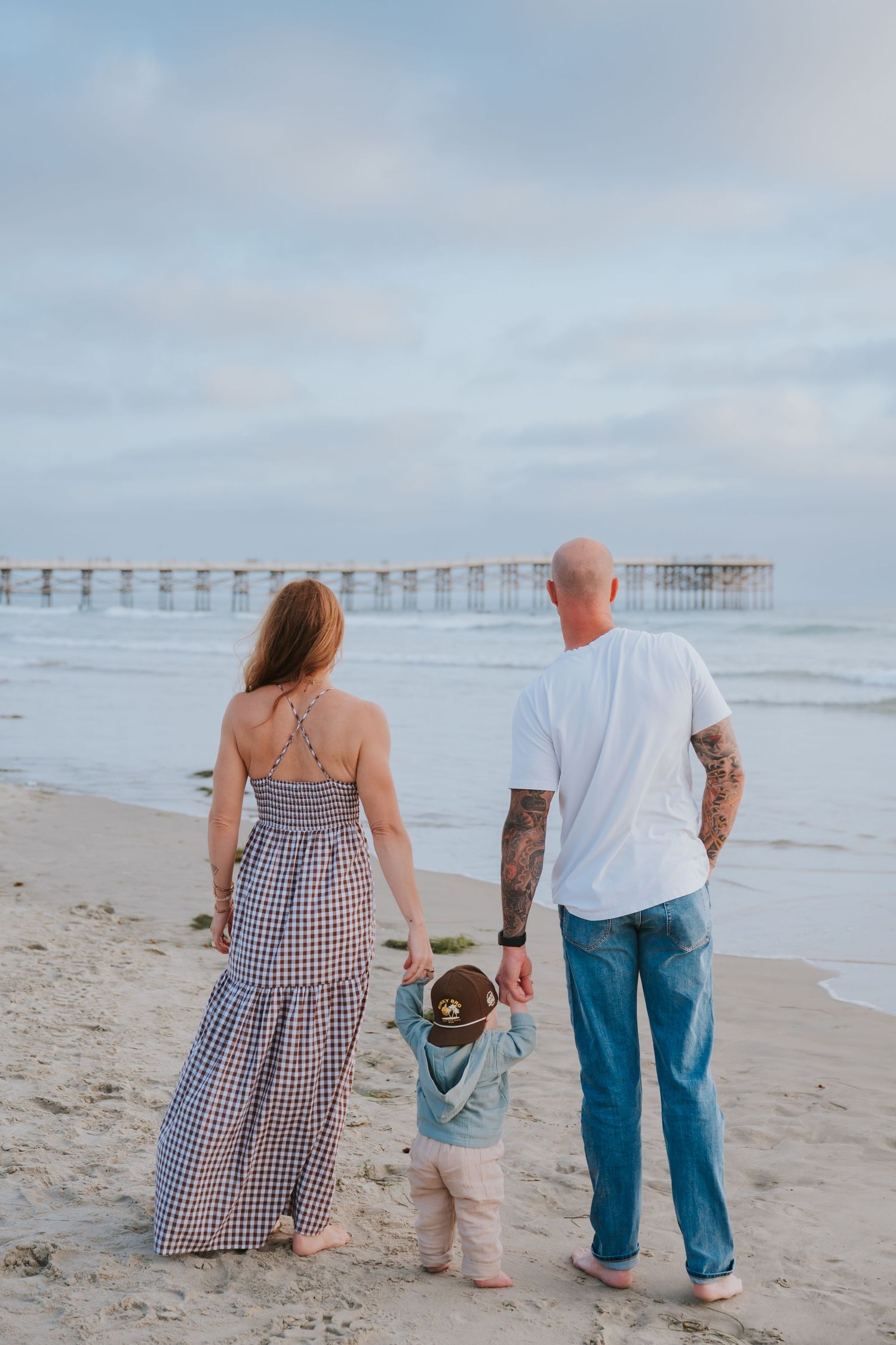 Family of three holding hands on sandy beach looking at ocean pier at dusk.